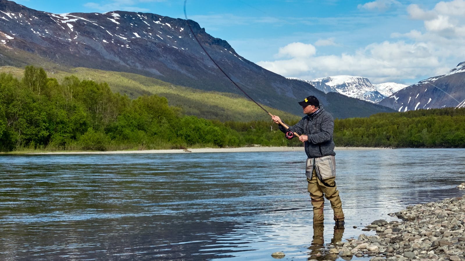 A man fly fishing in a river. There are mountains behind him and trees nearby. 