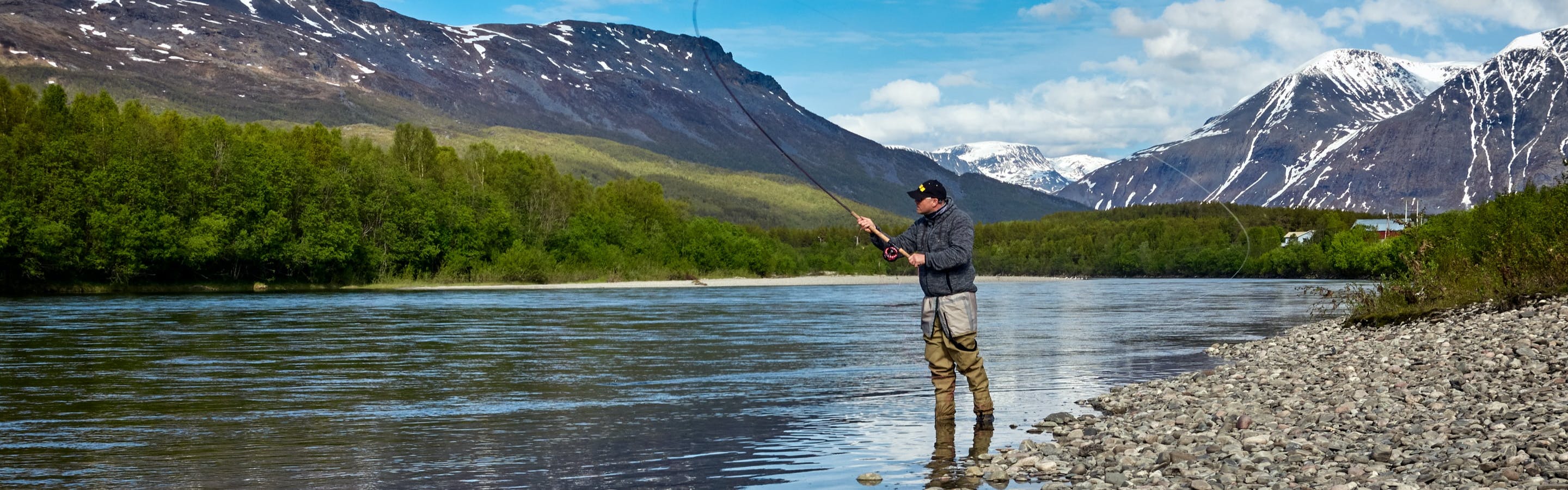 A man fly fishing in a river. There are mountains behind him and trees nearby. 