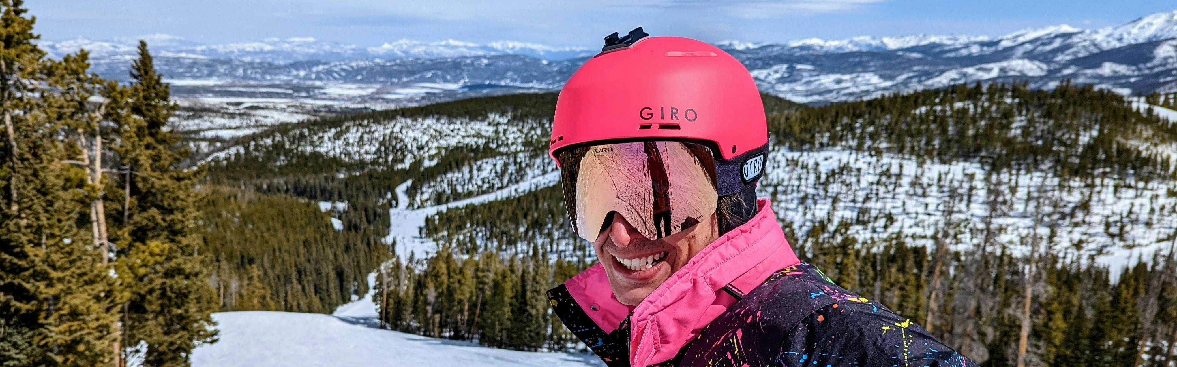 A skier smiling at the camera while wearing the Giro Emerge MIPS helmet. There is a ski run in the background. 