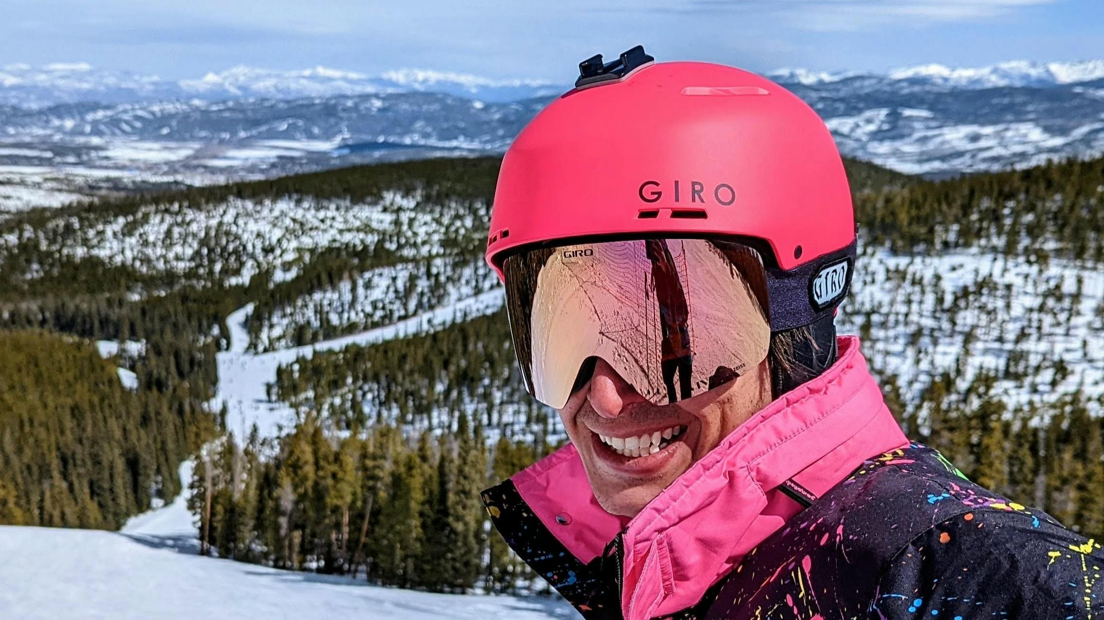 A skier smiling at the camera while wearing the Giro Emerge MIPS helmet. There is a ski run in the background. 