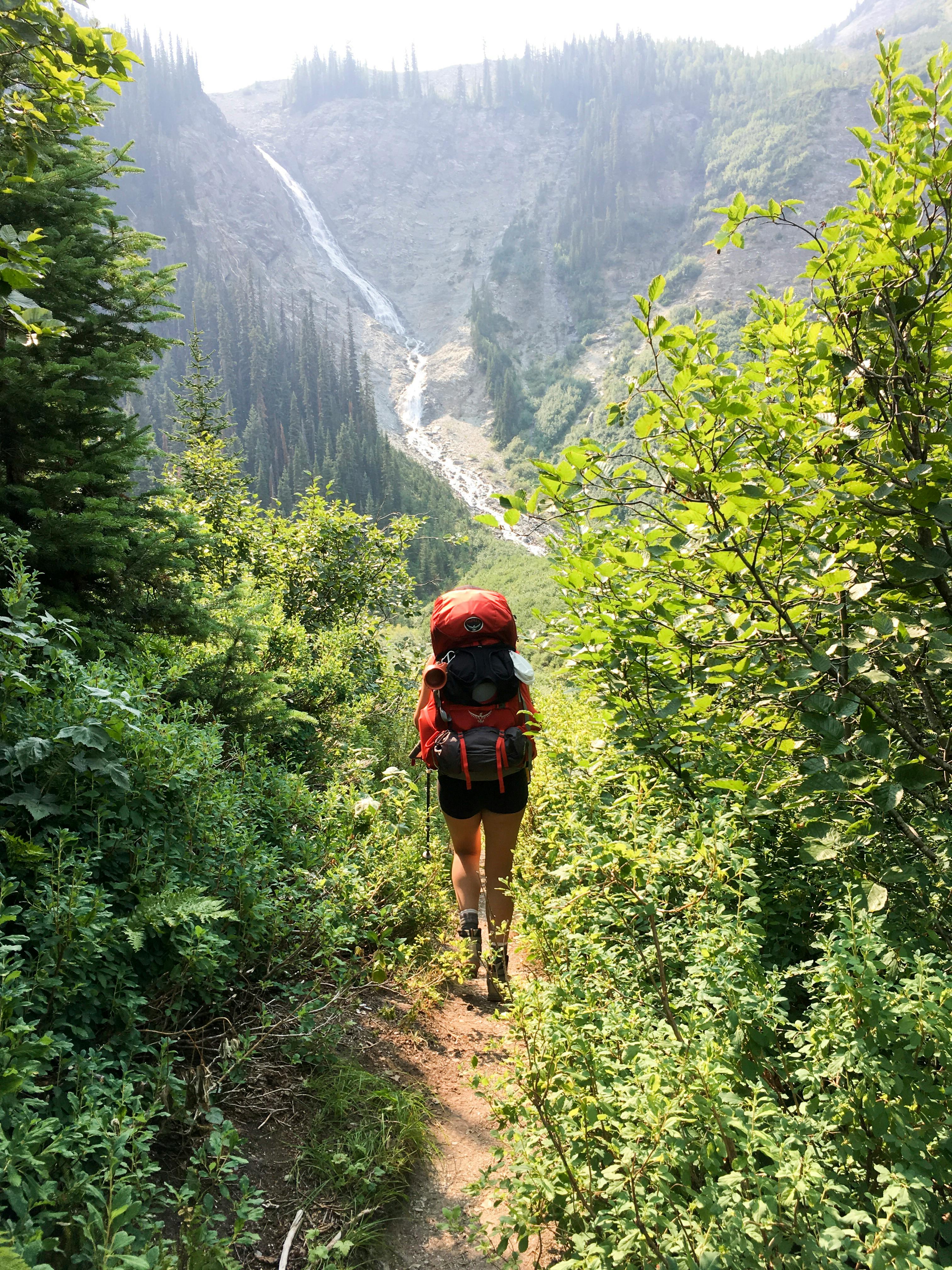 A woman with a red backpacking pack walks away from the camera down a narrow trail lined with trees