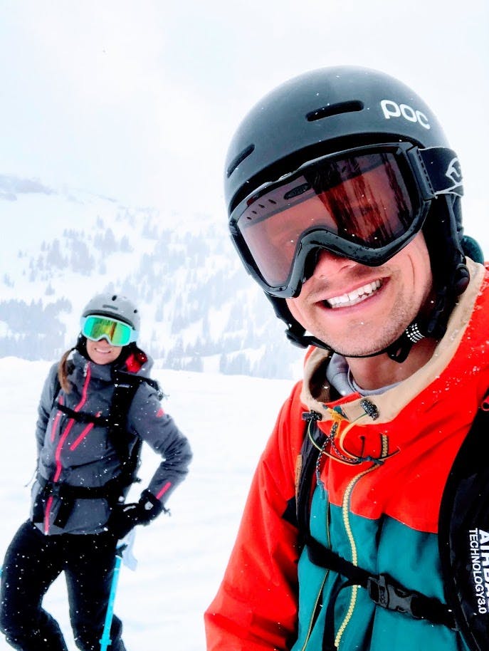 A man takes a selfie with the author behind him. They are in helmets, goggles, ski apparel, and wear backpacks. The ground is heavily snowy, and the visibility is poor. 