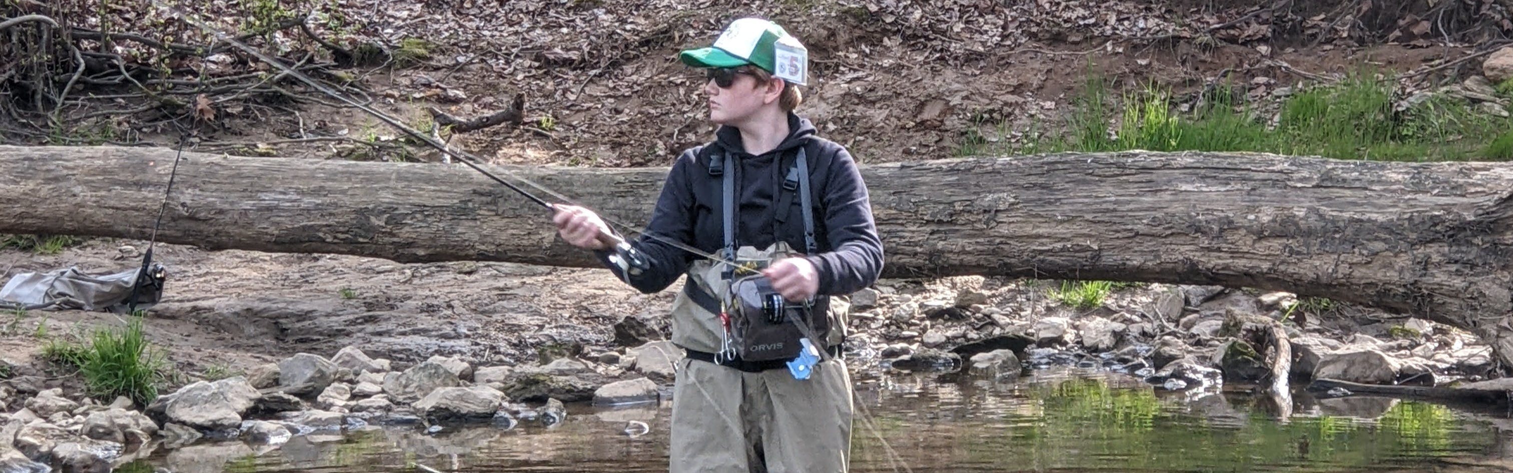 A boy casts a fly fishing rod into a river. He is wearing waders and a hat.