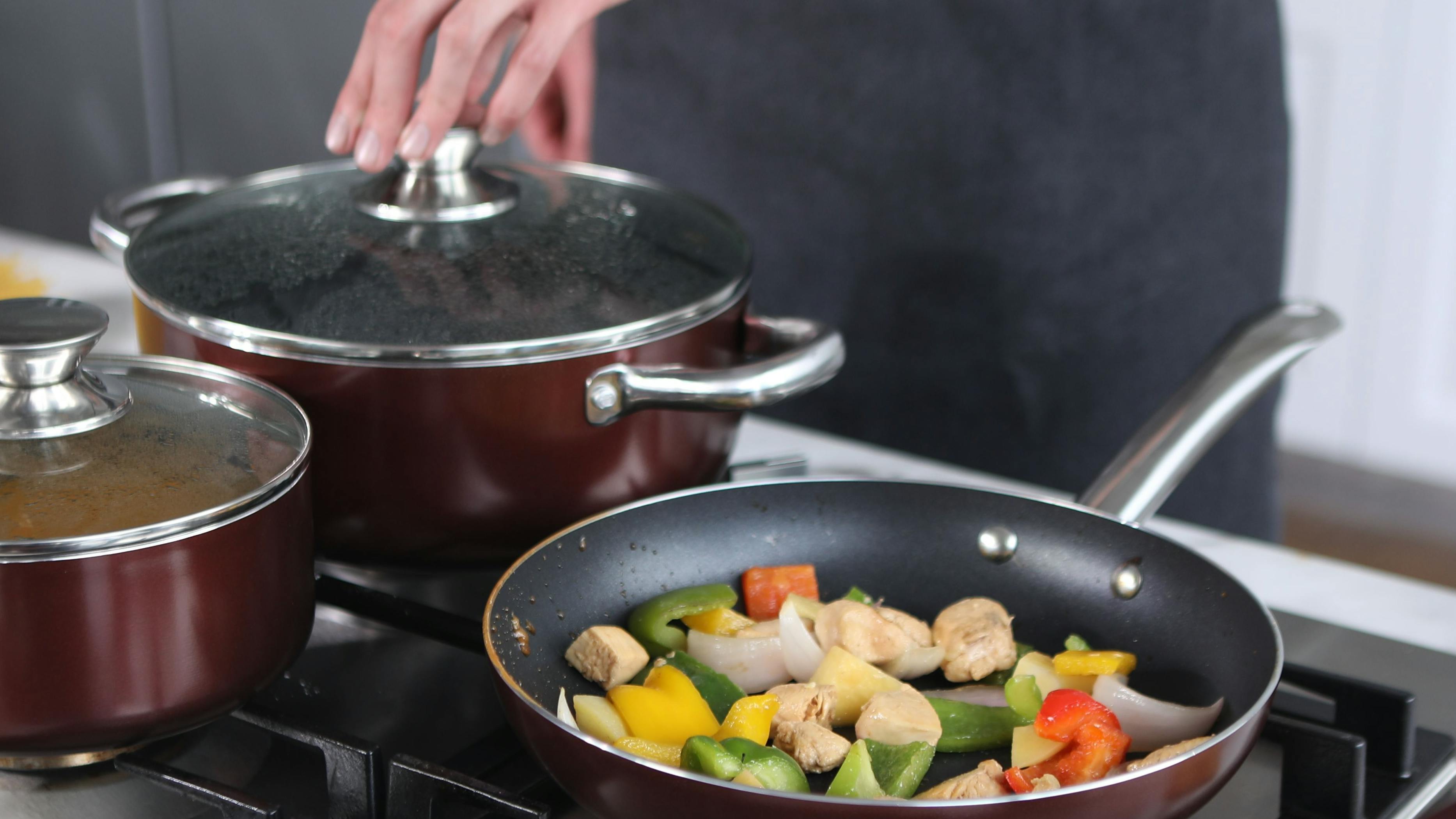 Someone uses a matching cookware set over a stove to make some spaghetti and fry some vegetables. 