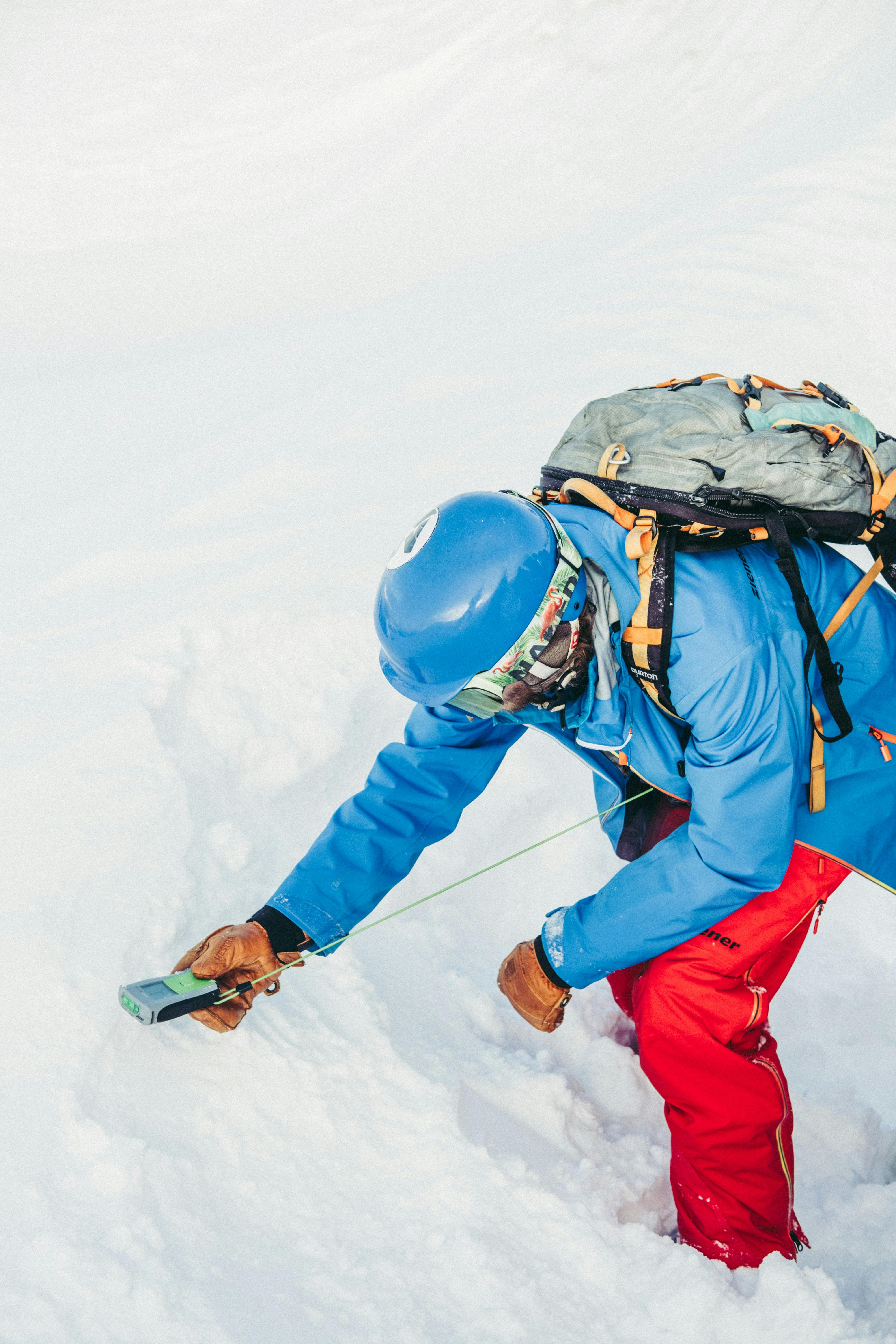 A person in a blue jacket and red pants uses an avalanche beacon