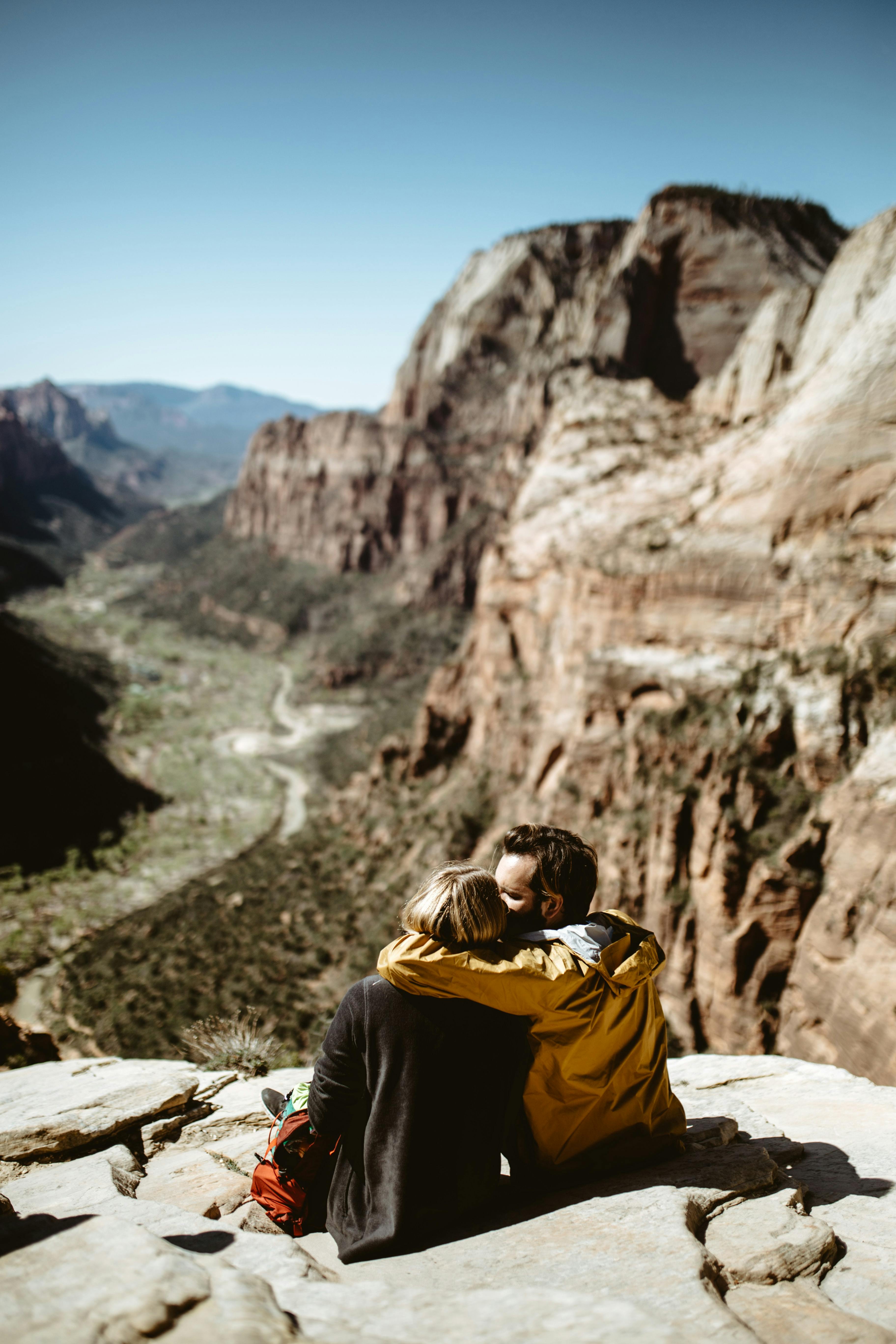 A man and a woman sitting at the top of a hike. The man has his arm around the woman and is kissing her on the cheek.