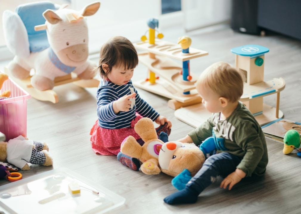 Two toddlers playing with a teddy bear surrounded by toys