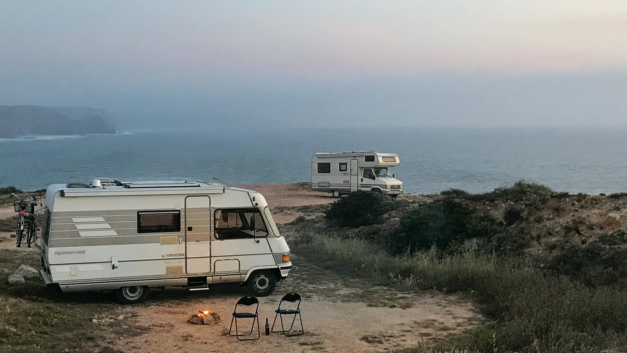 Two RVs are parked on an overlook along the coast. Next to one RV are two folding chairs and a campfire. 