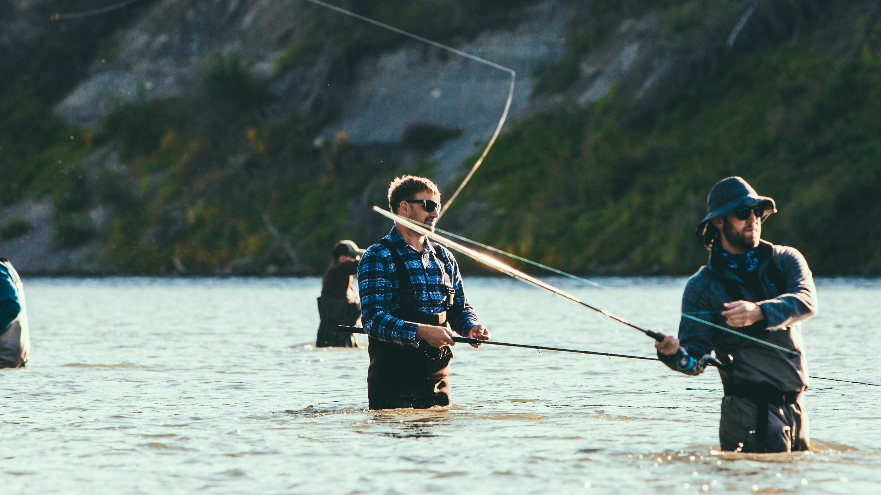 Six men fly fishing in a river, all spread out and talking amongst themselves. 