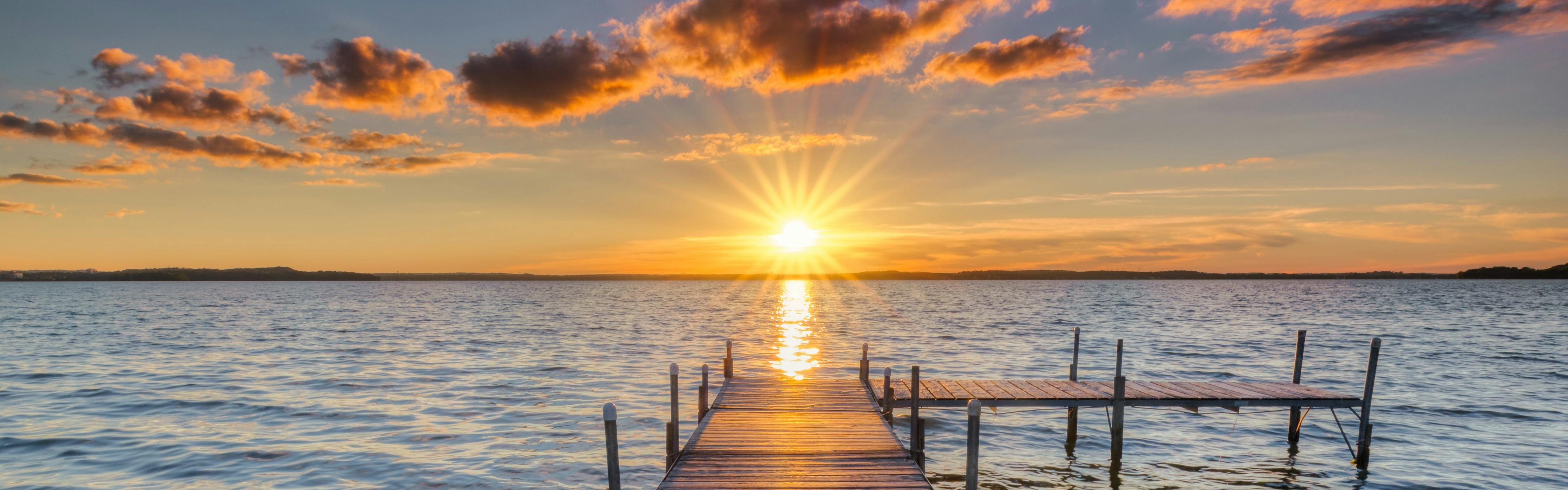 A dock takes an L-turn to the right in front of the setting sun which is sending orange right out in rays.