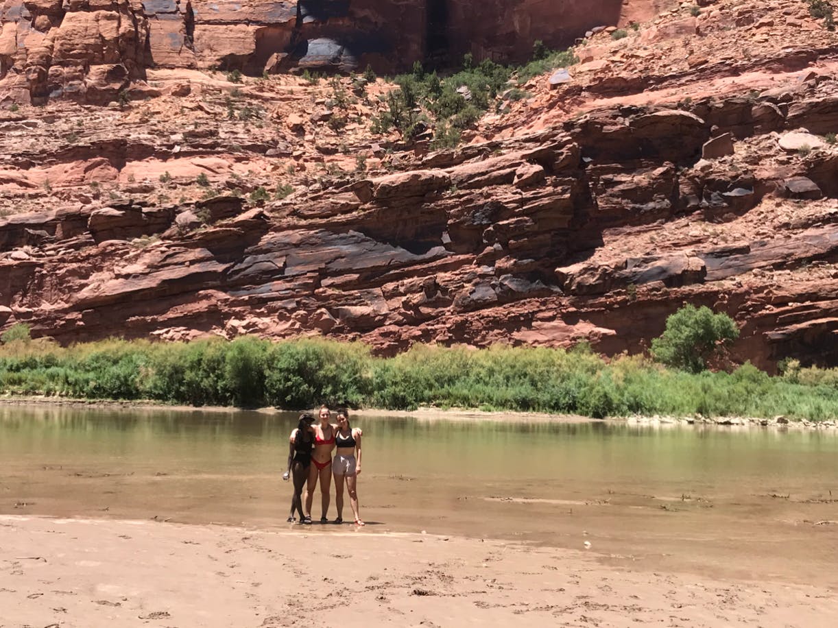 Three girls stand with their feet in the water of a shallow river. The mud and background canyon walls are red. 