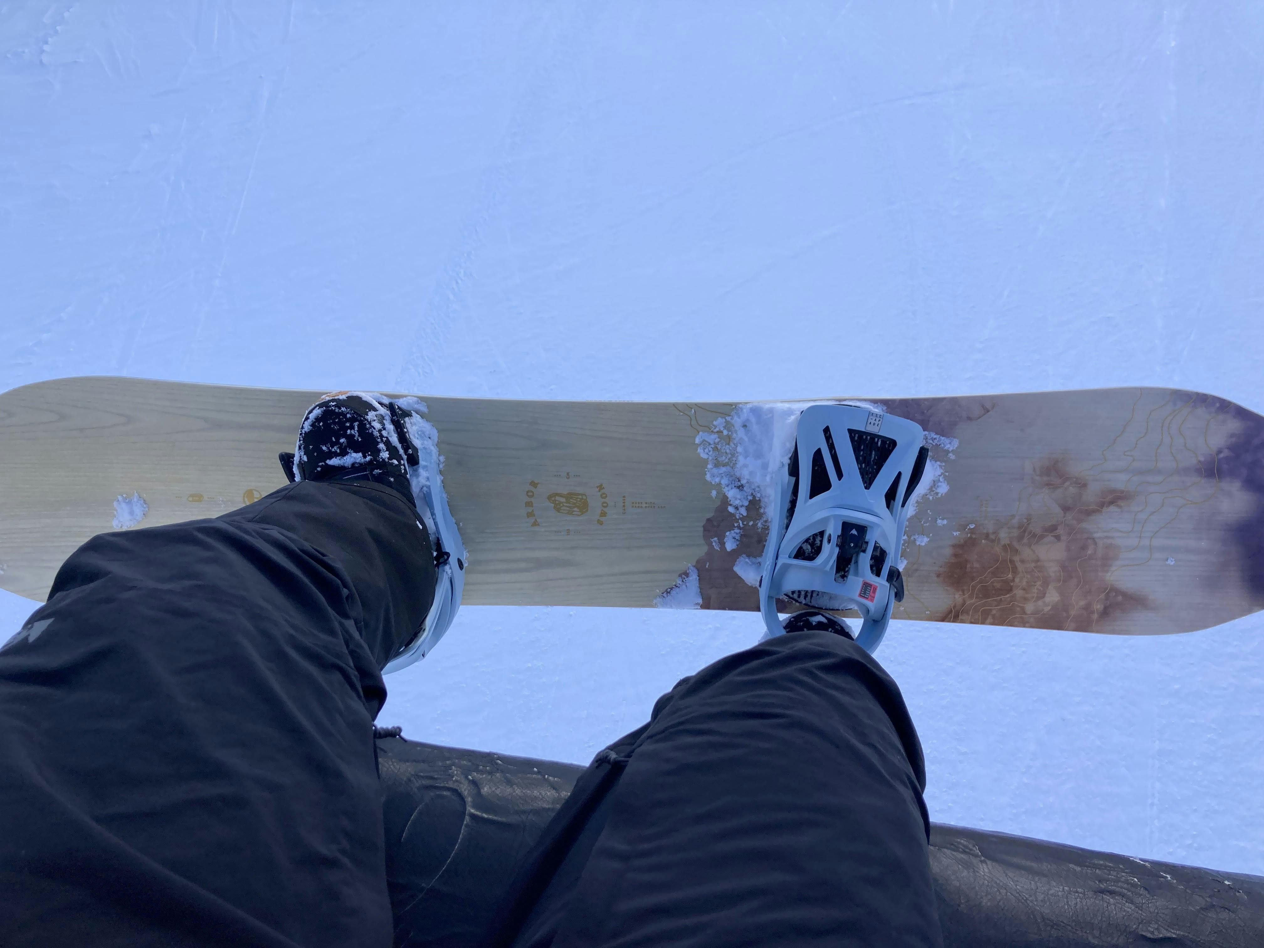Looking down at a snowboarder's board, feet and bindings while riding up the lift. The left foot is strapped in, while the right is unstrapped.