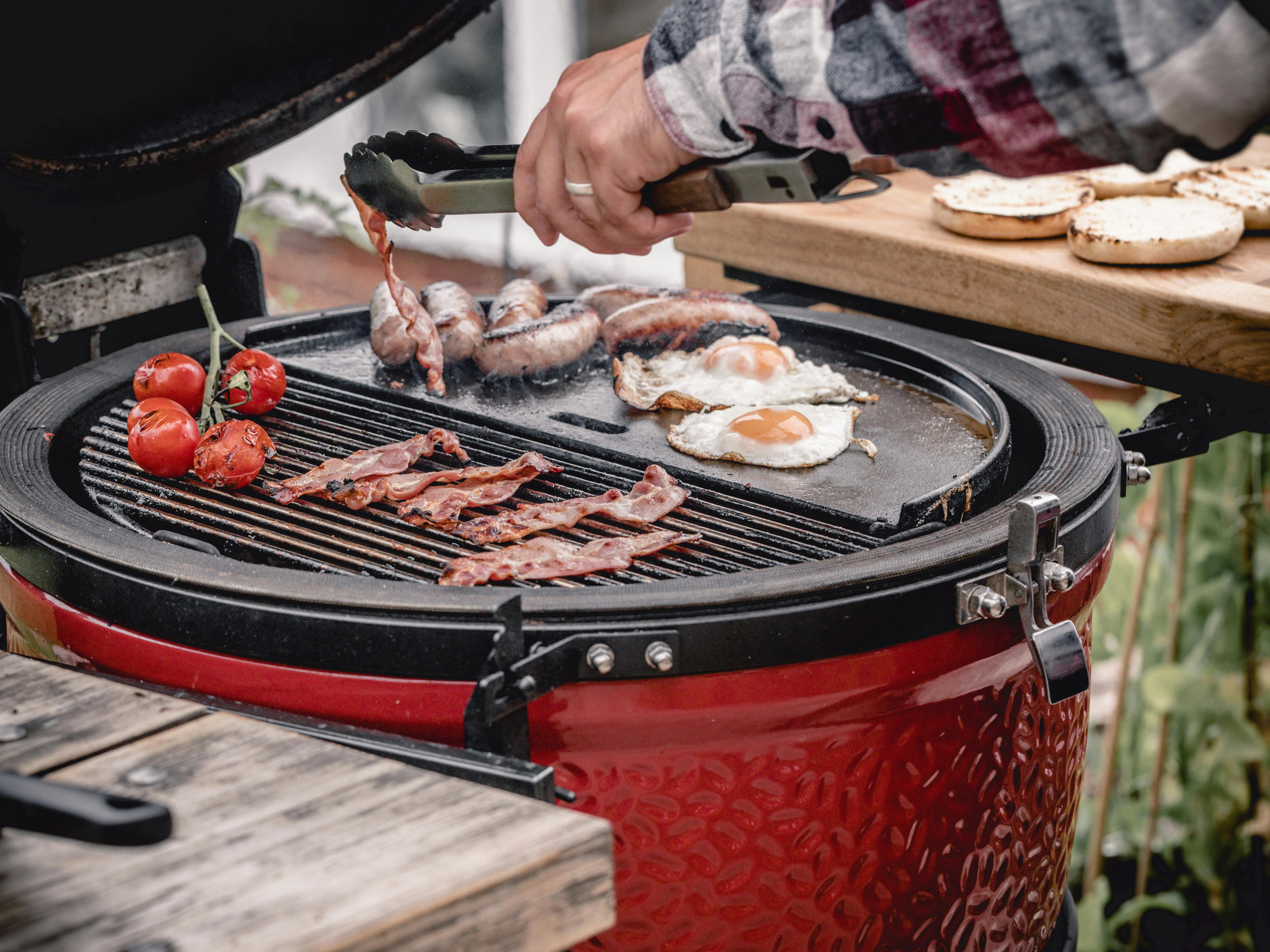 A grill master moves some bacon on a Kamado Grill. There are also some eggs and tomatoes on the grill. 