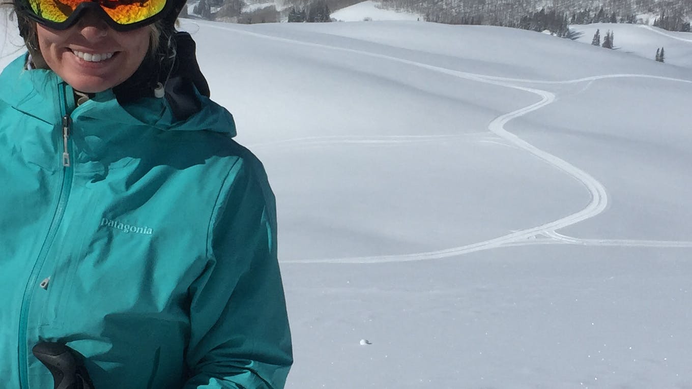 A woman in a ski jacket. There is a ski track behind her and a lot of snow. She is wearing goggles. 