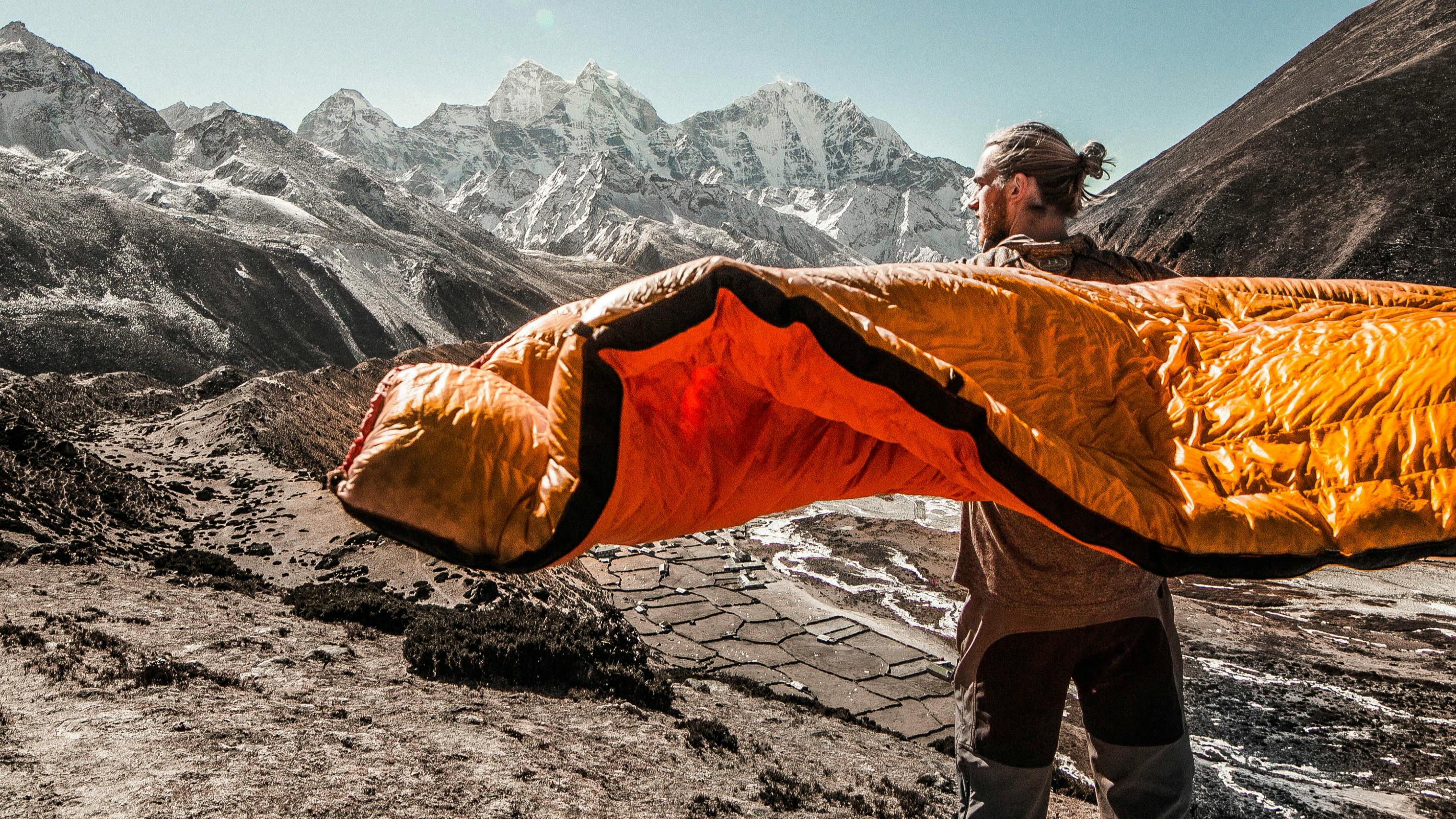 A man stretches out an orange sleeping bag over his shoulders and it catches in the wind. 