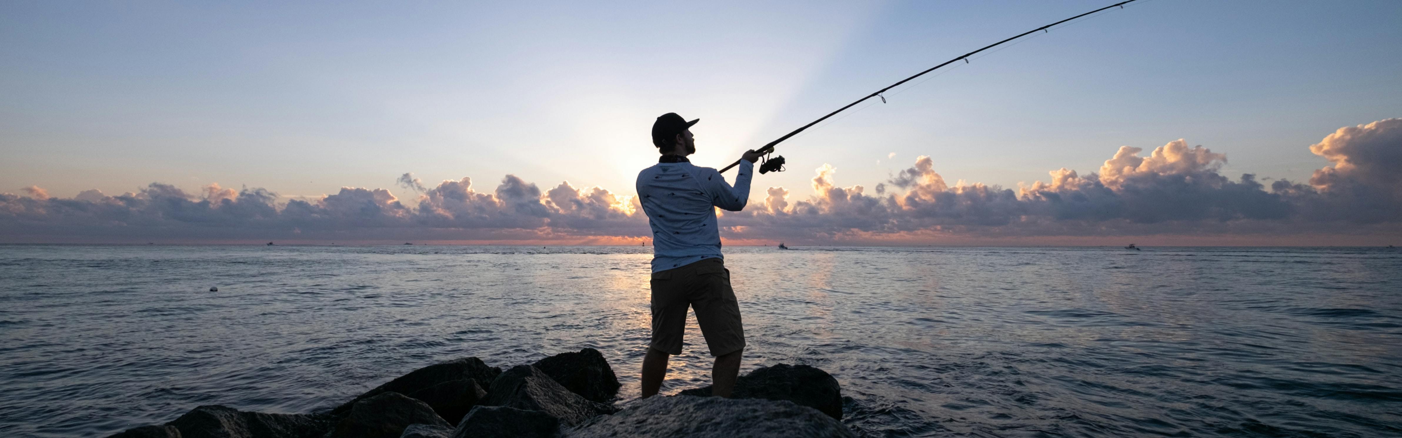 A man fishes off of a jetty with the sun rising in front of him.