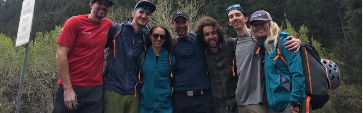 Seven people stand with outdoor gear hugging. 