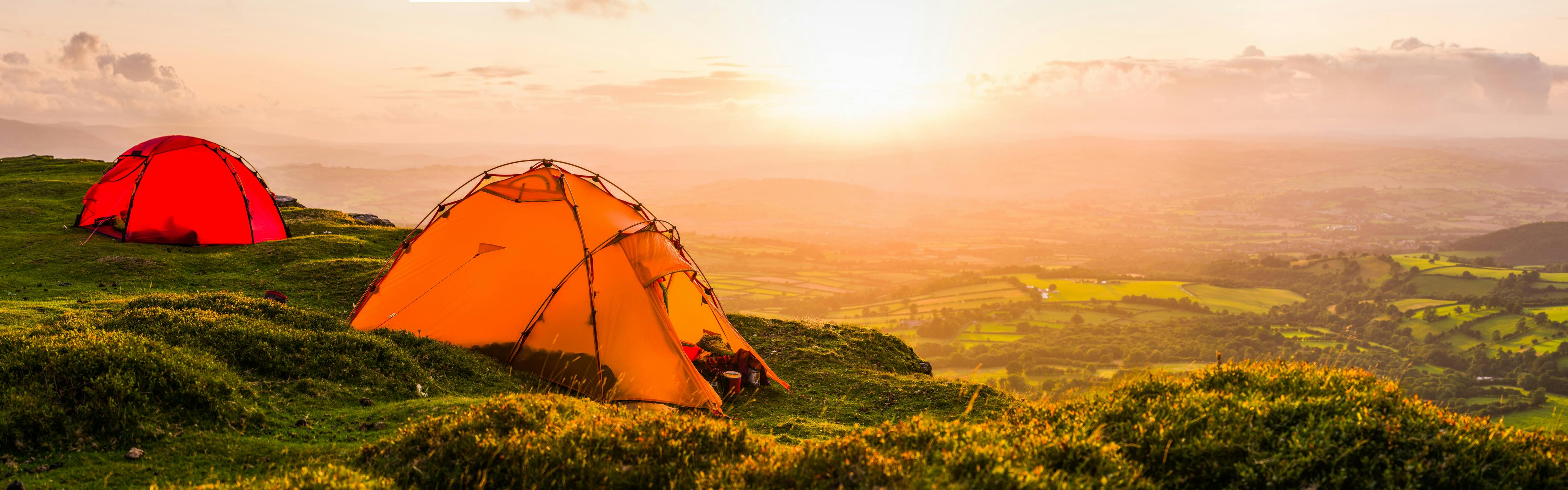 Two tents - one orange and one red - sit on a grassy hill as the sun rises