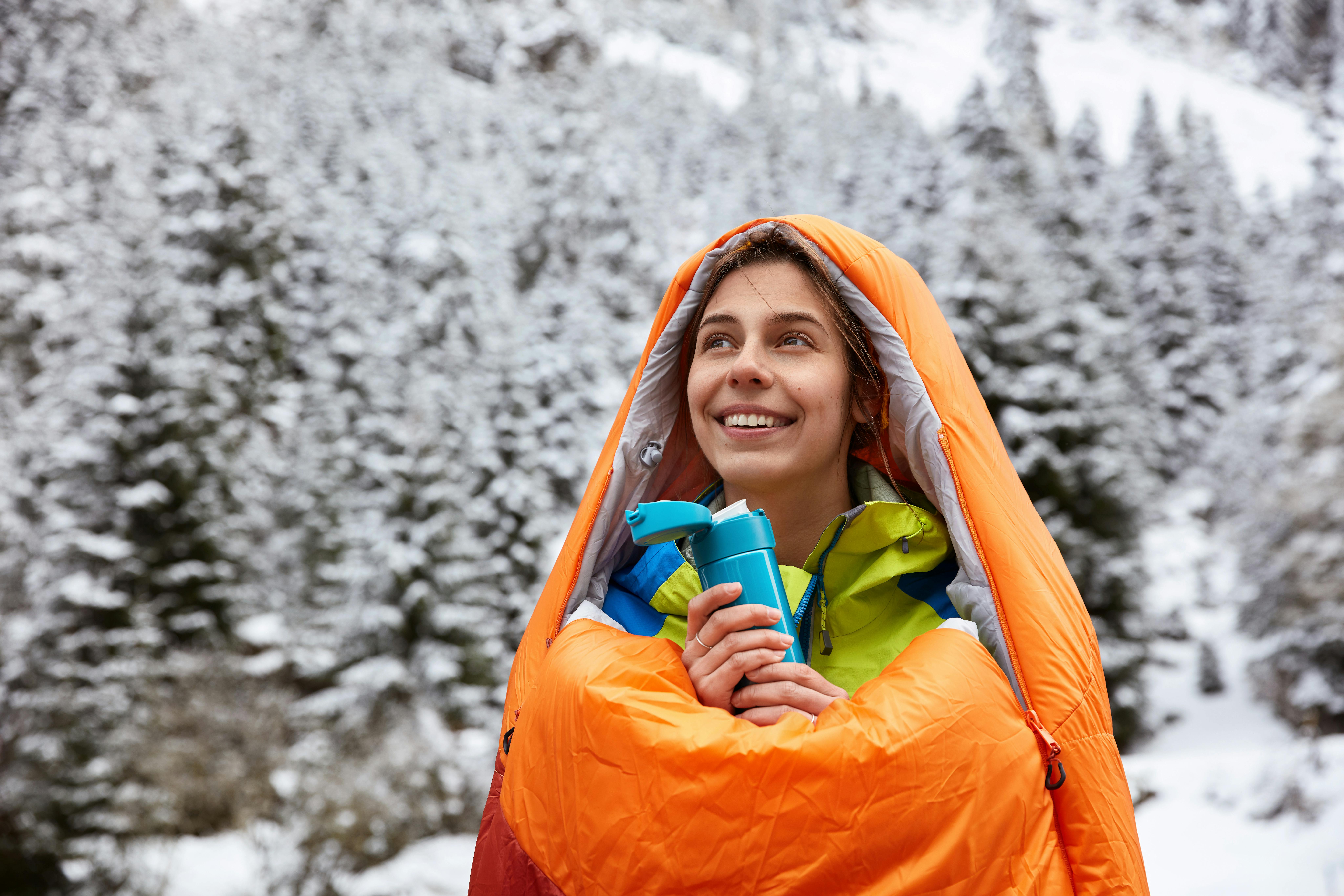 A woman in a sleeping bag. There is a snow and snowy trees behind her. 