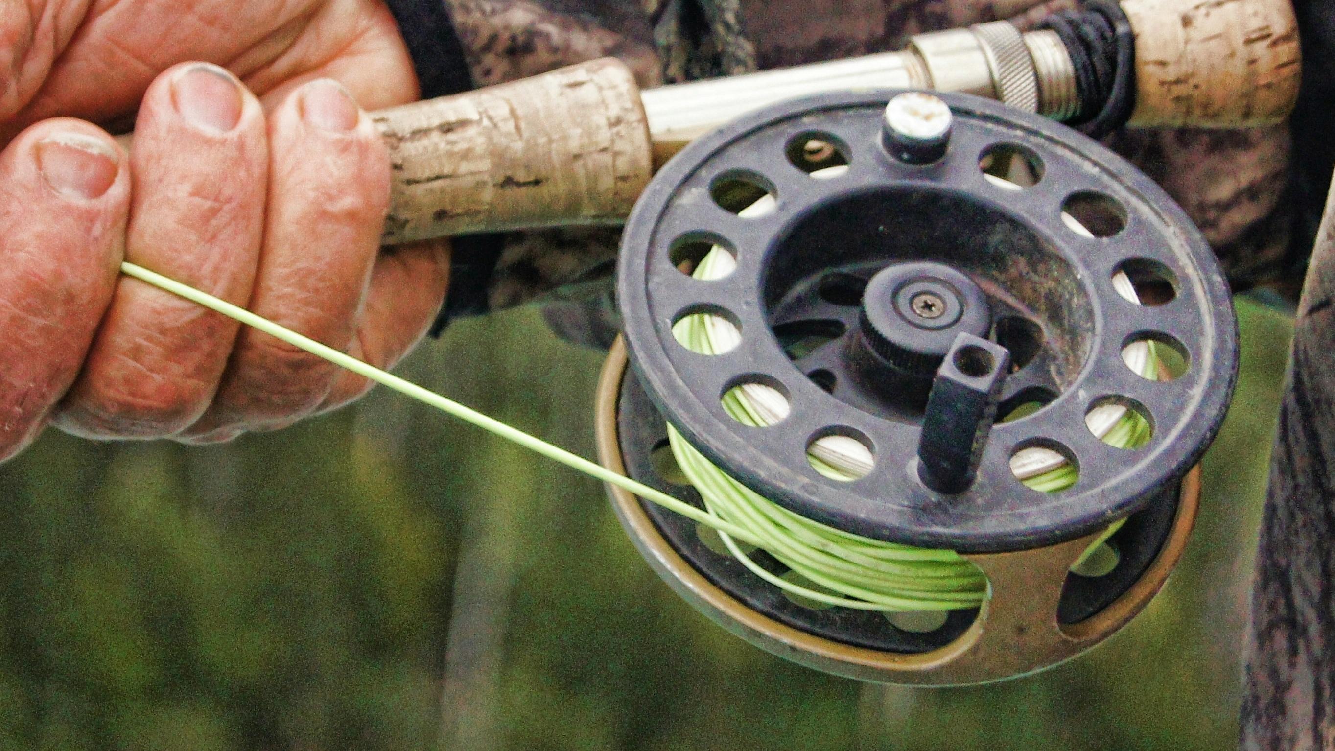 A closeup of a man with rough hands holding a flyfishing rod centered on the reel which he holds close to his body. 