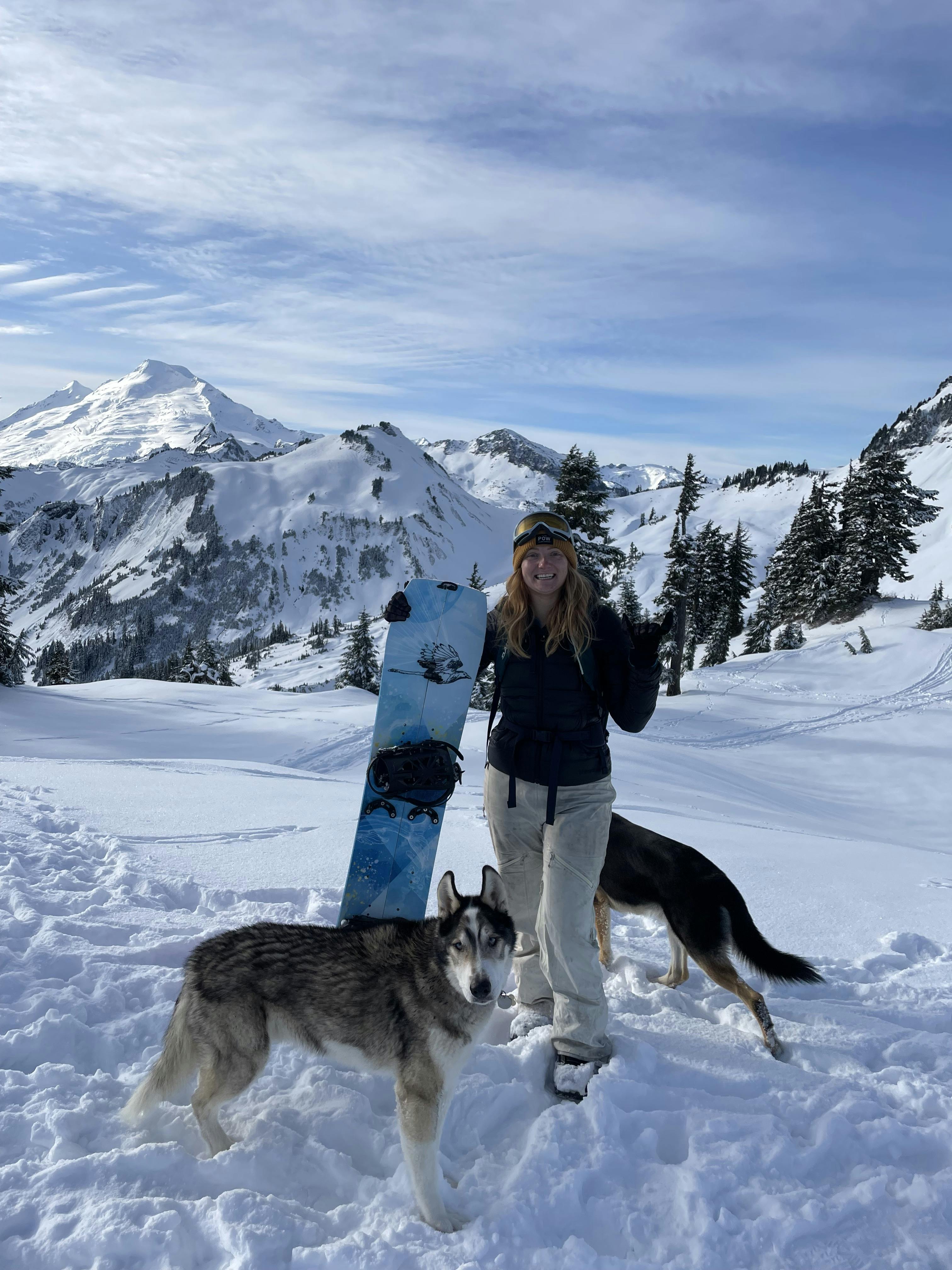 A woman poses in a snowy area with two dogs and her snowboard. There are snowy mountains in the background.