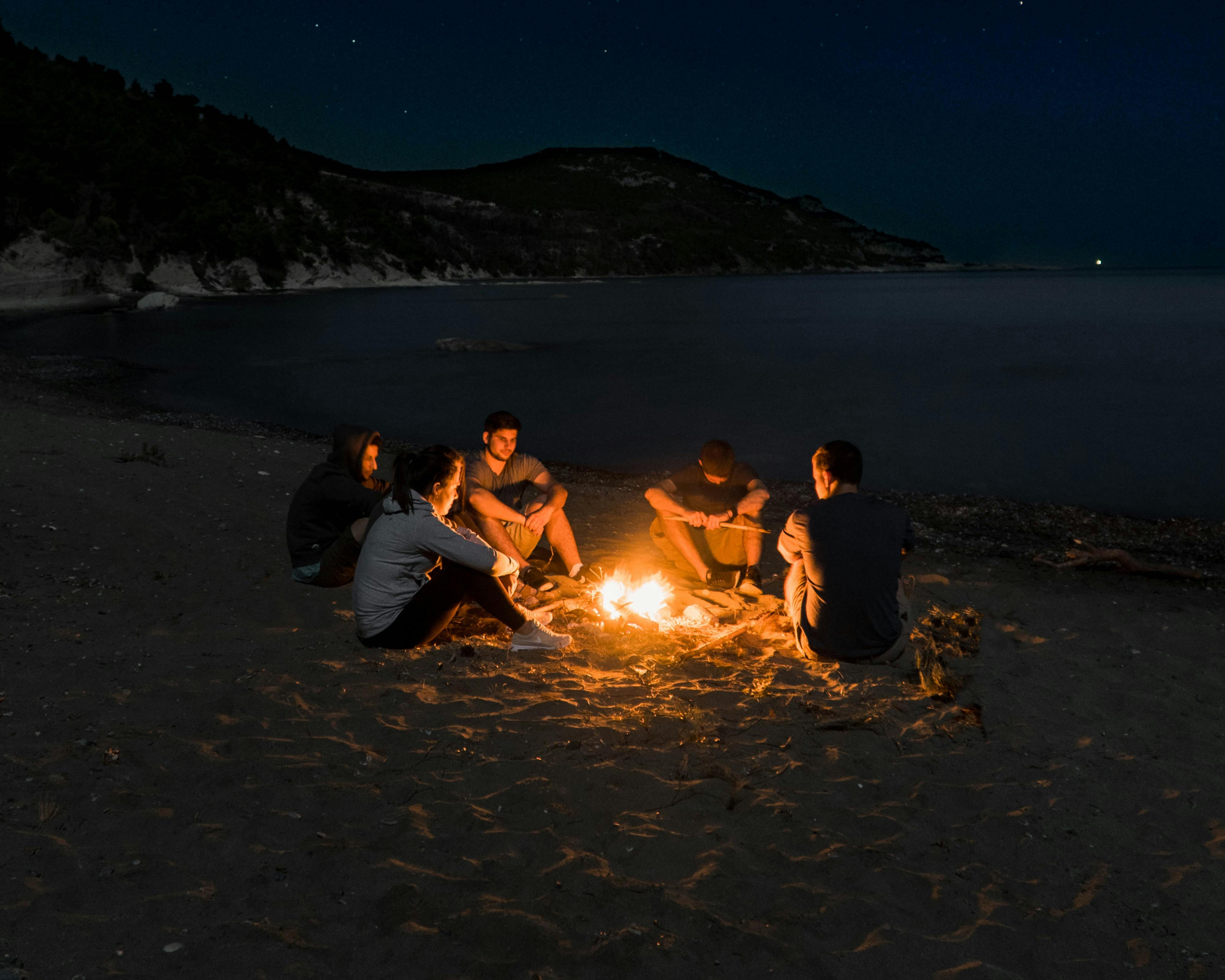 A group of people sit around a campfire on a beach at night