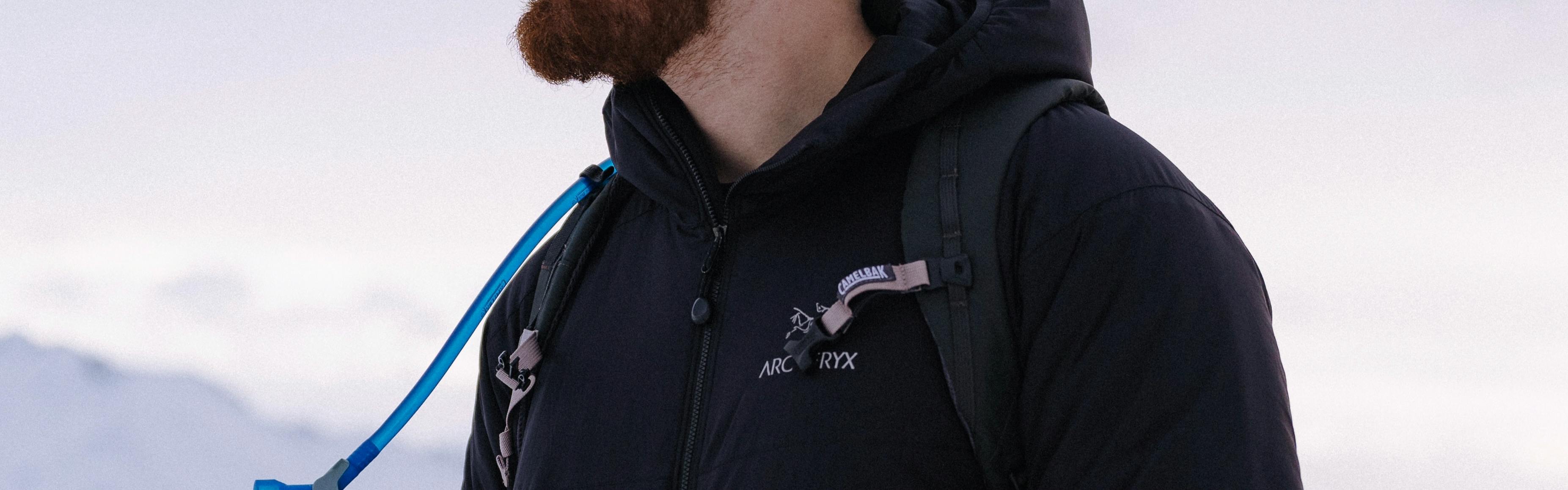 Close up of a man wearing a jacket. Just his chest and beard is visible. He has a backpack on and you can see mountains in the  background.