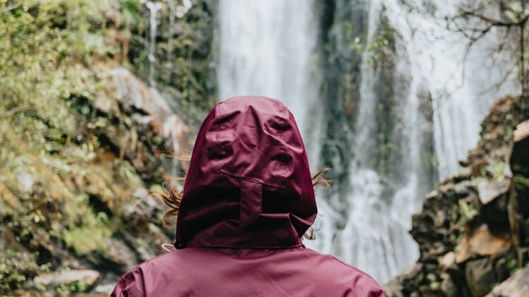 Someone in a maroon raincoat stands in front of a waterfall. 