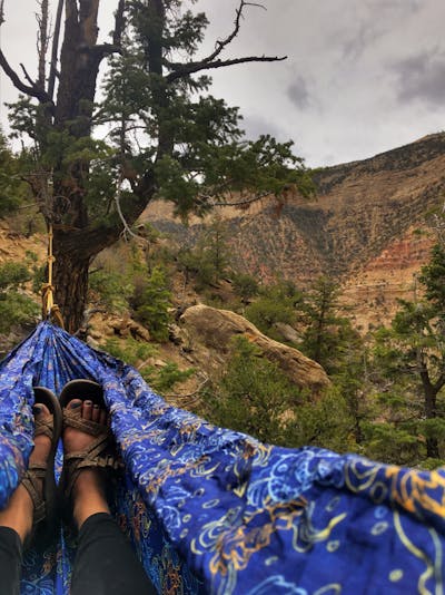 Feet in a hammock wearing sandals. There is a tree and mountains visible in the distance. 