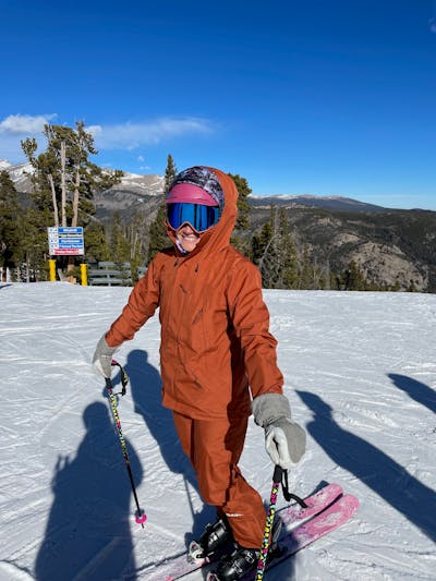 A woman wearing an orange. jacket and pants stands on her pink skis on a ski run.