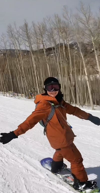 A woman snowboards down a mountain wearing orange pants and an orange jacket. She smiles at the camera as she goes by.