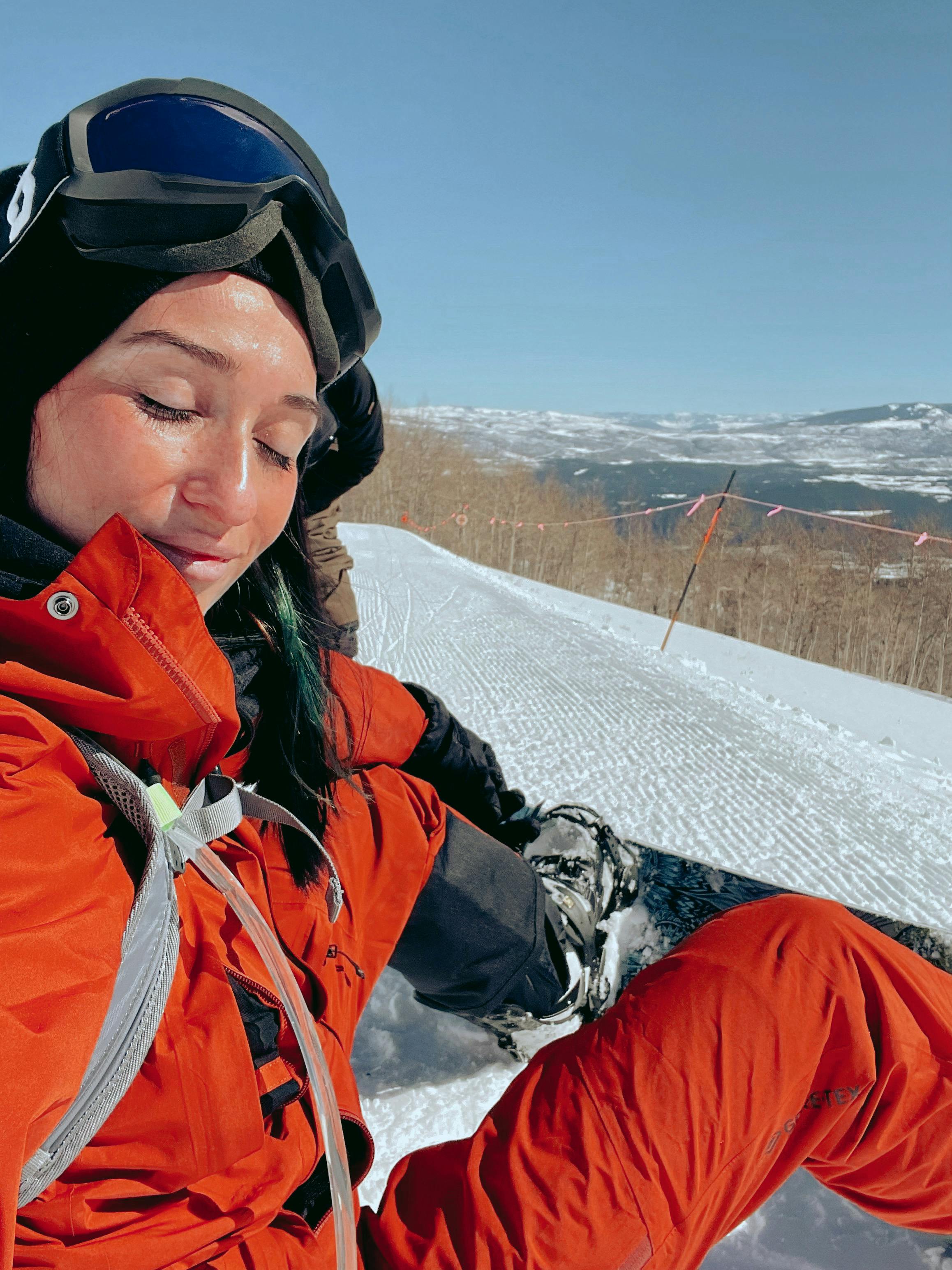 A woman in an orange outfit wearing a snowboard on her feet smiles at the camera. She is sitting on a hill at a ski resort.