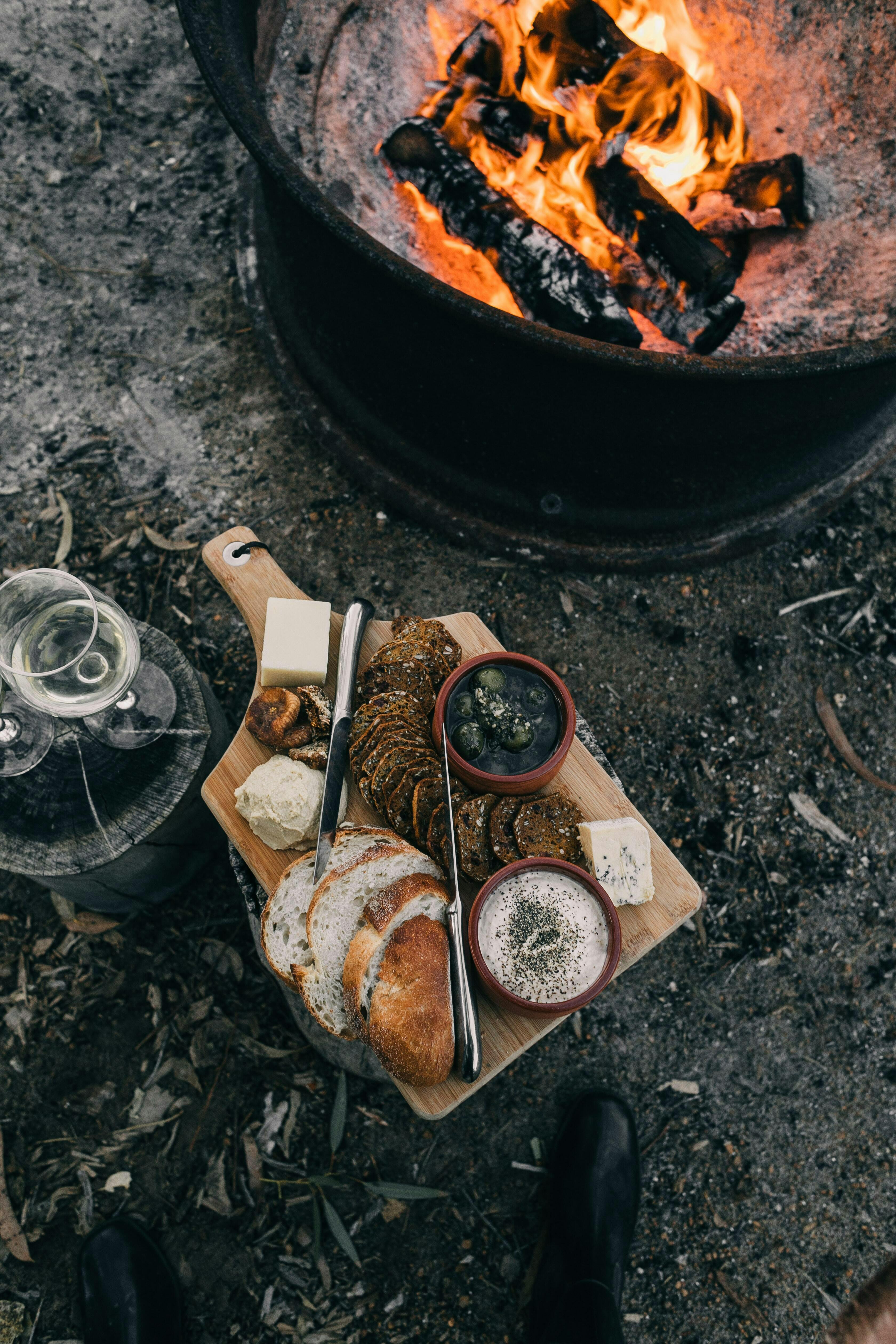 A charcuterie board sits near a fire pit. There are a few glasses of wine on the side and bread, olives, cheese, and hummus on the board.