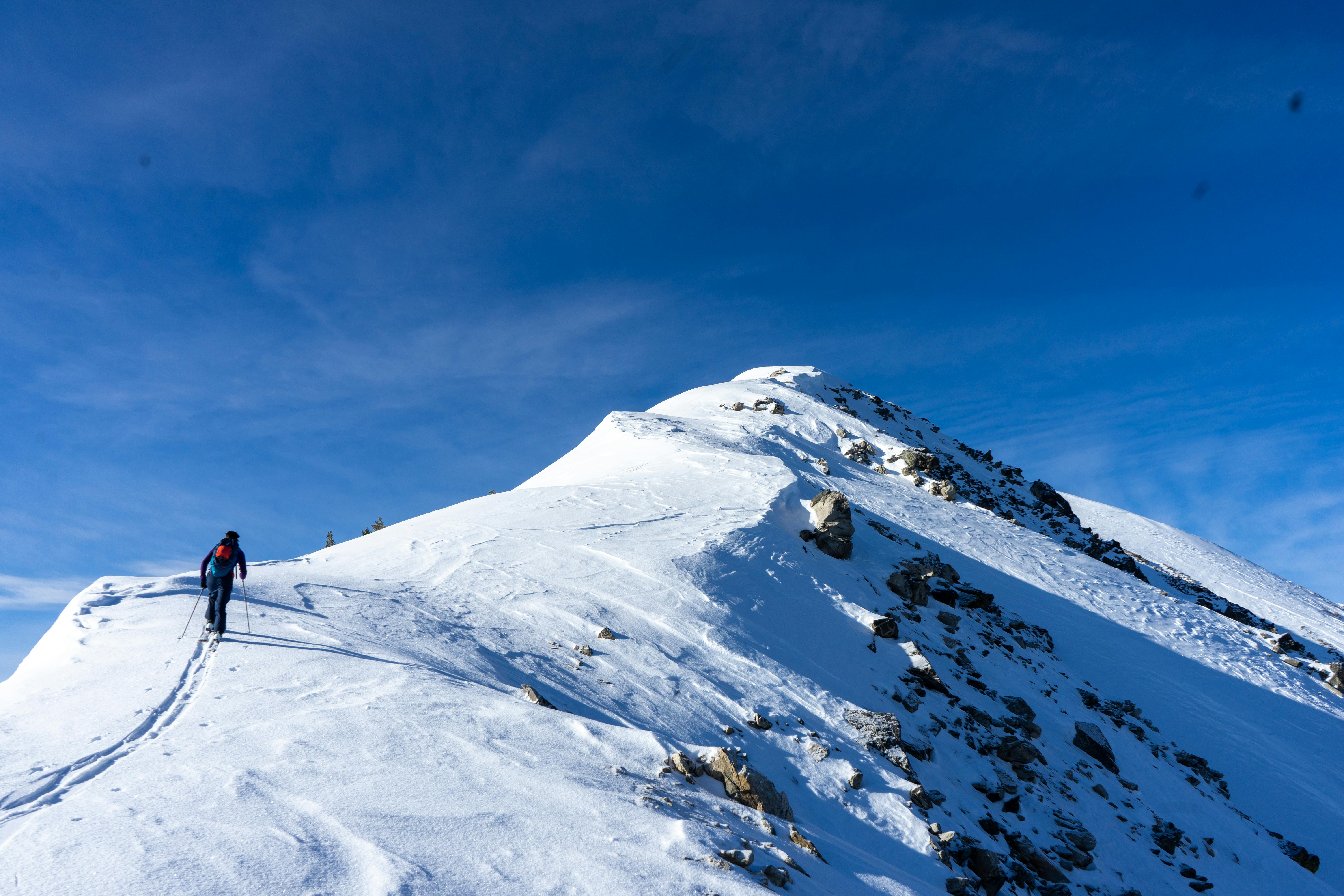 A skier ascends a mountain to ski. He is almost at the top and the sky is very blue. There is a lot of snow and a lot of rocks.