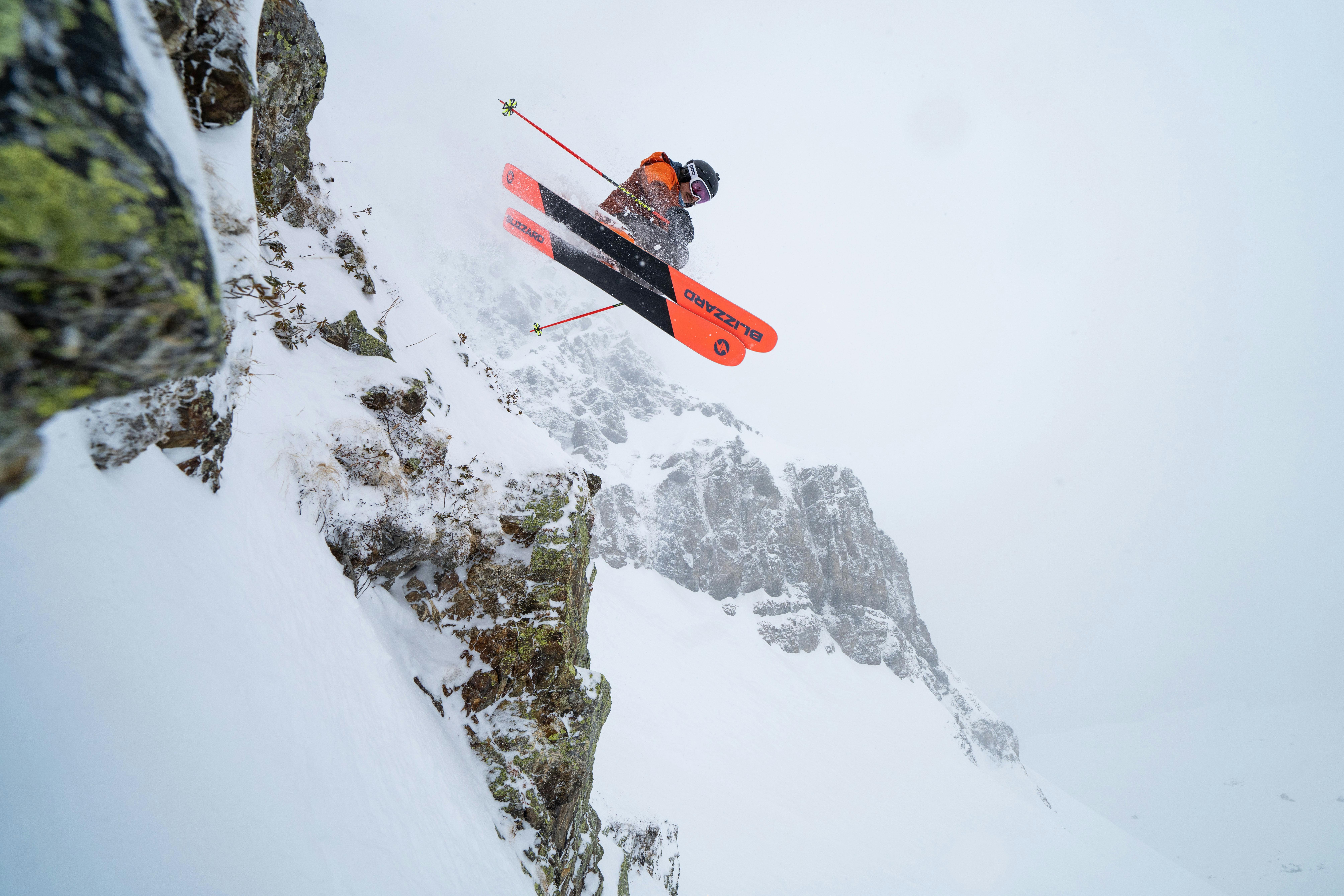 A skier jumping off a snowy cliff with bright orange and black Blizzard skis