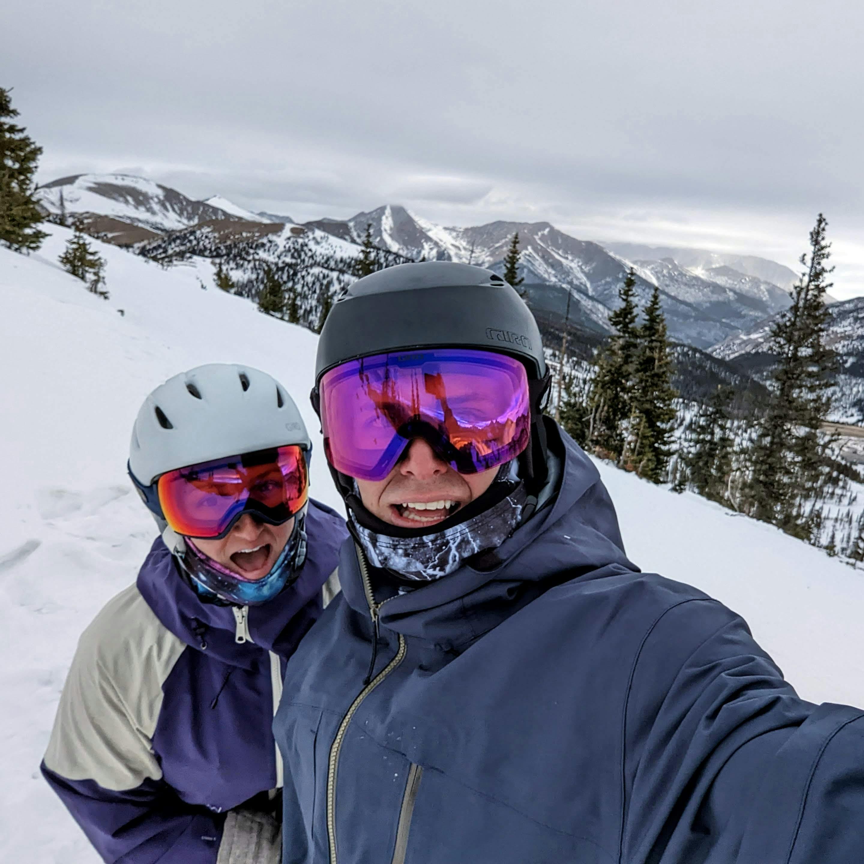 Selfie of two skiers on a ski run. There are snowy mountains in the background. 