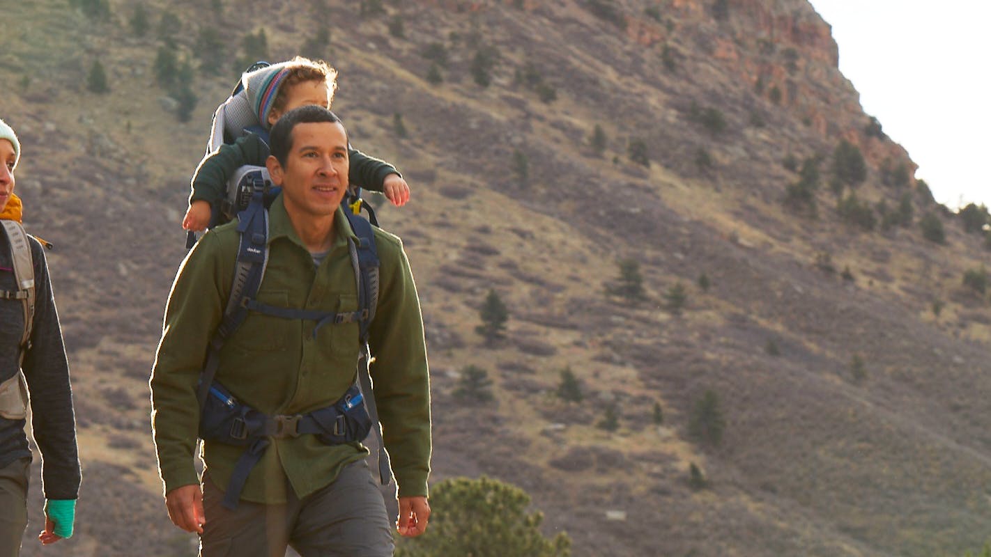 A man walks with a Deuter backpack and child carrier on his back that a toddler is sitting in. A woman walks next to them.
