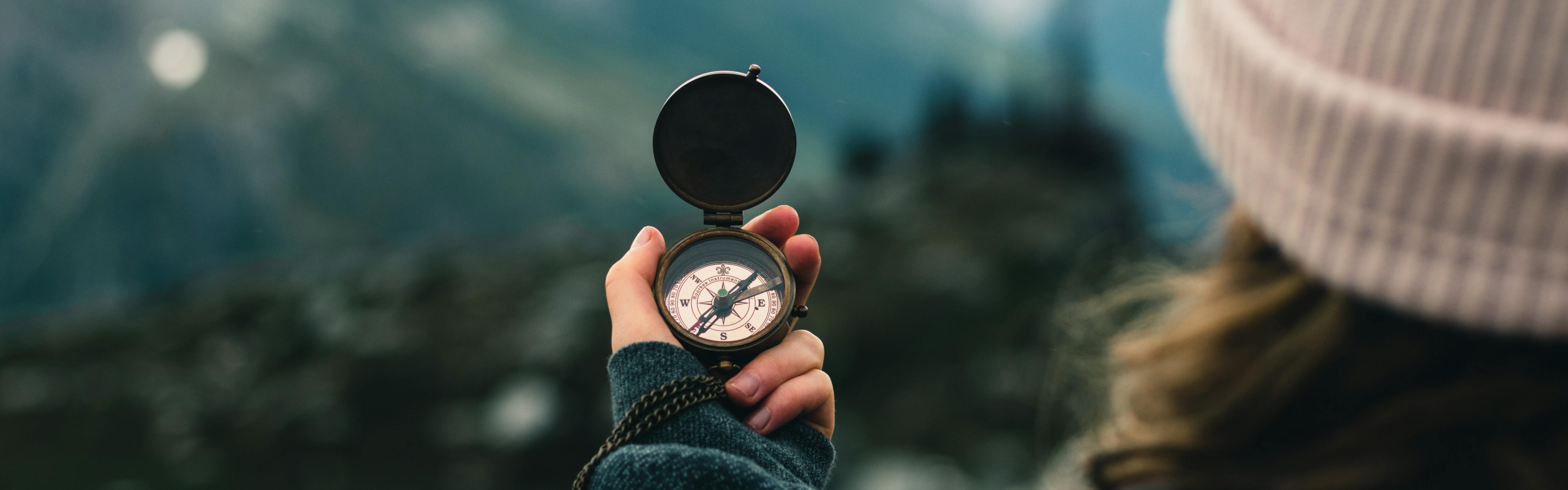 A woman in a beanie holds open a compass in front of a green landscape and we see the image from behind her.  