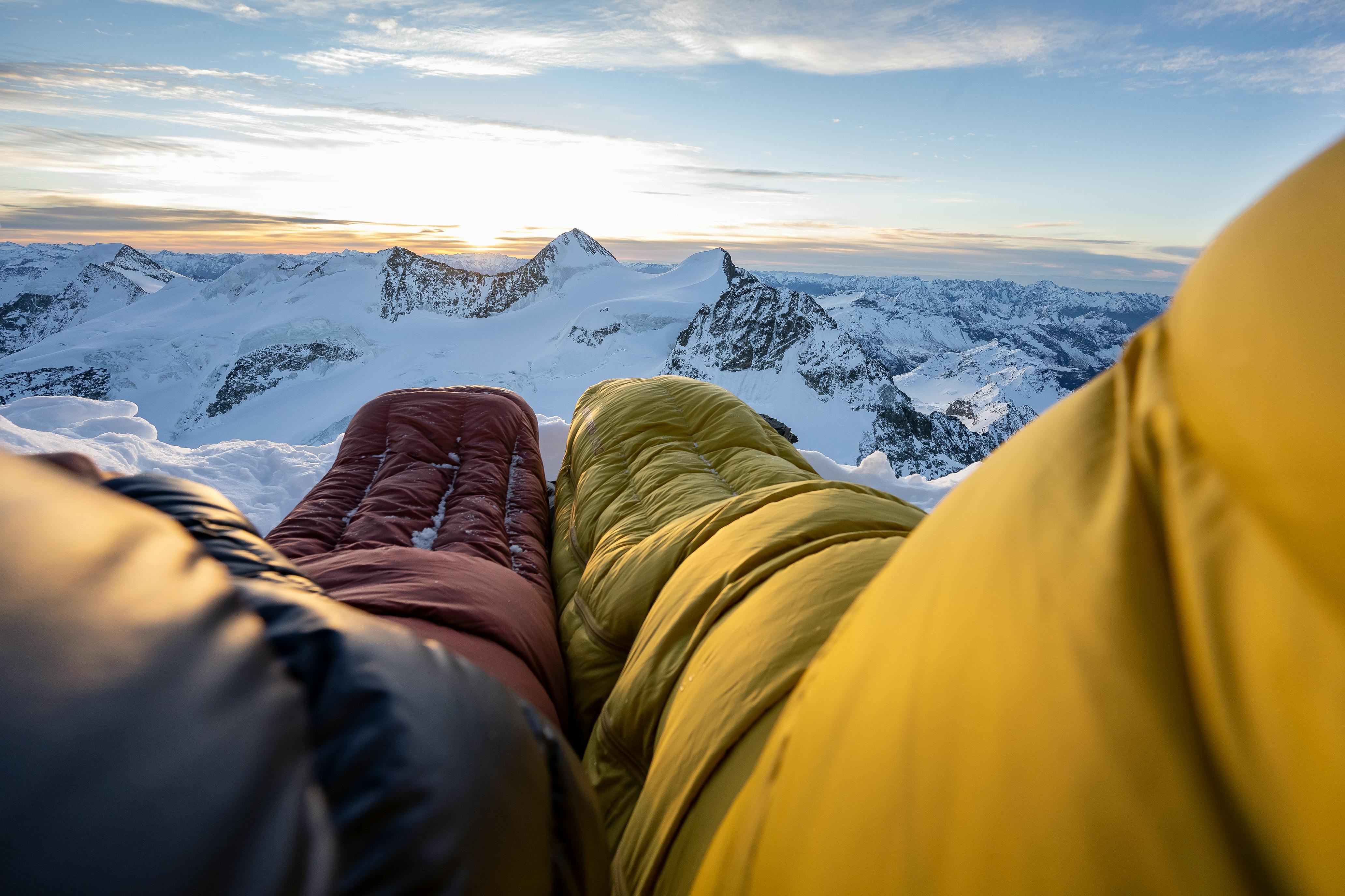 Looking down at two sleeping bags, a yellow and red one, and out at the snowy sunset mountain scenery beyond.