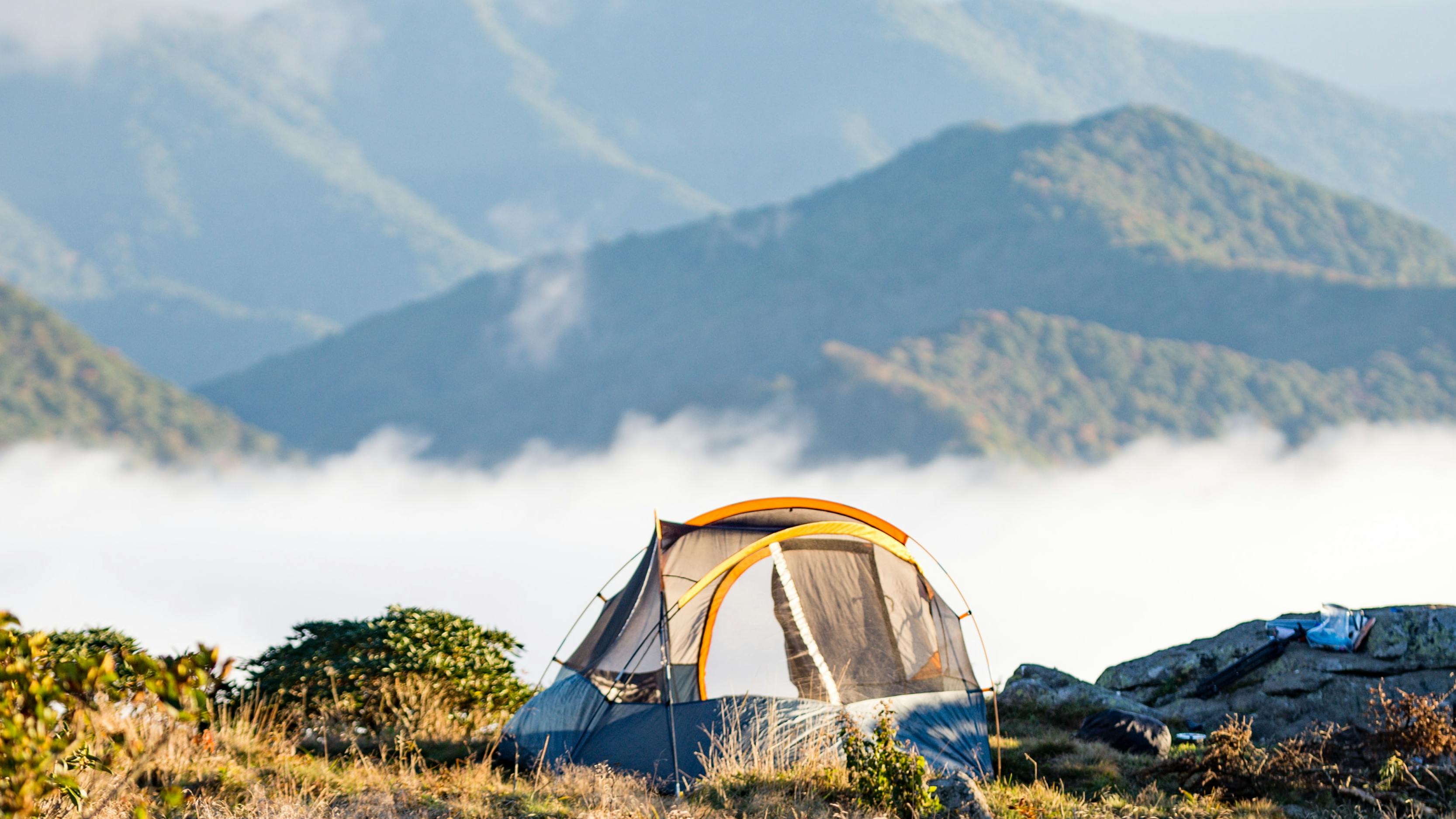 A tent is perched on a hillside, overlooked green, ridged mountains in the background. Fog fills the valley in between. 
