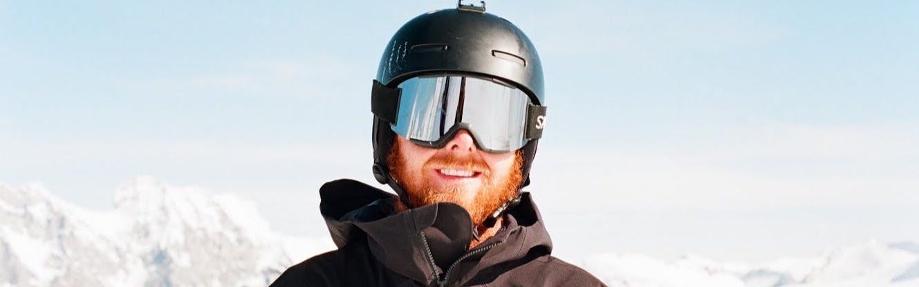 A man wearing the Smith Squad goggles on a sunny day with the ChromaPop Sun Platinum Mirror lens. There is a snowy mountain range behind him.