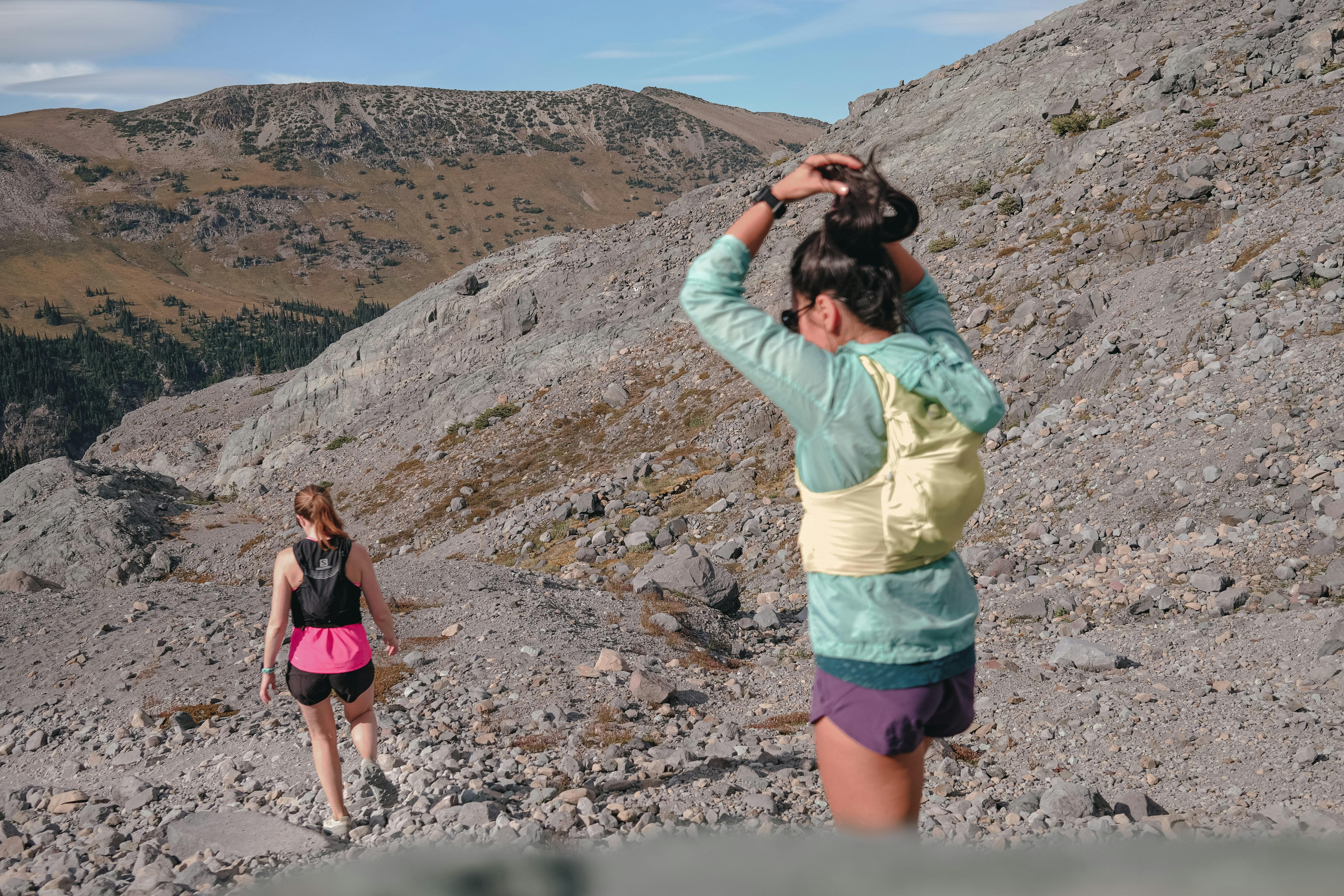 Two women with trail running vests on are on a trail with mountains in the background.
