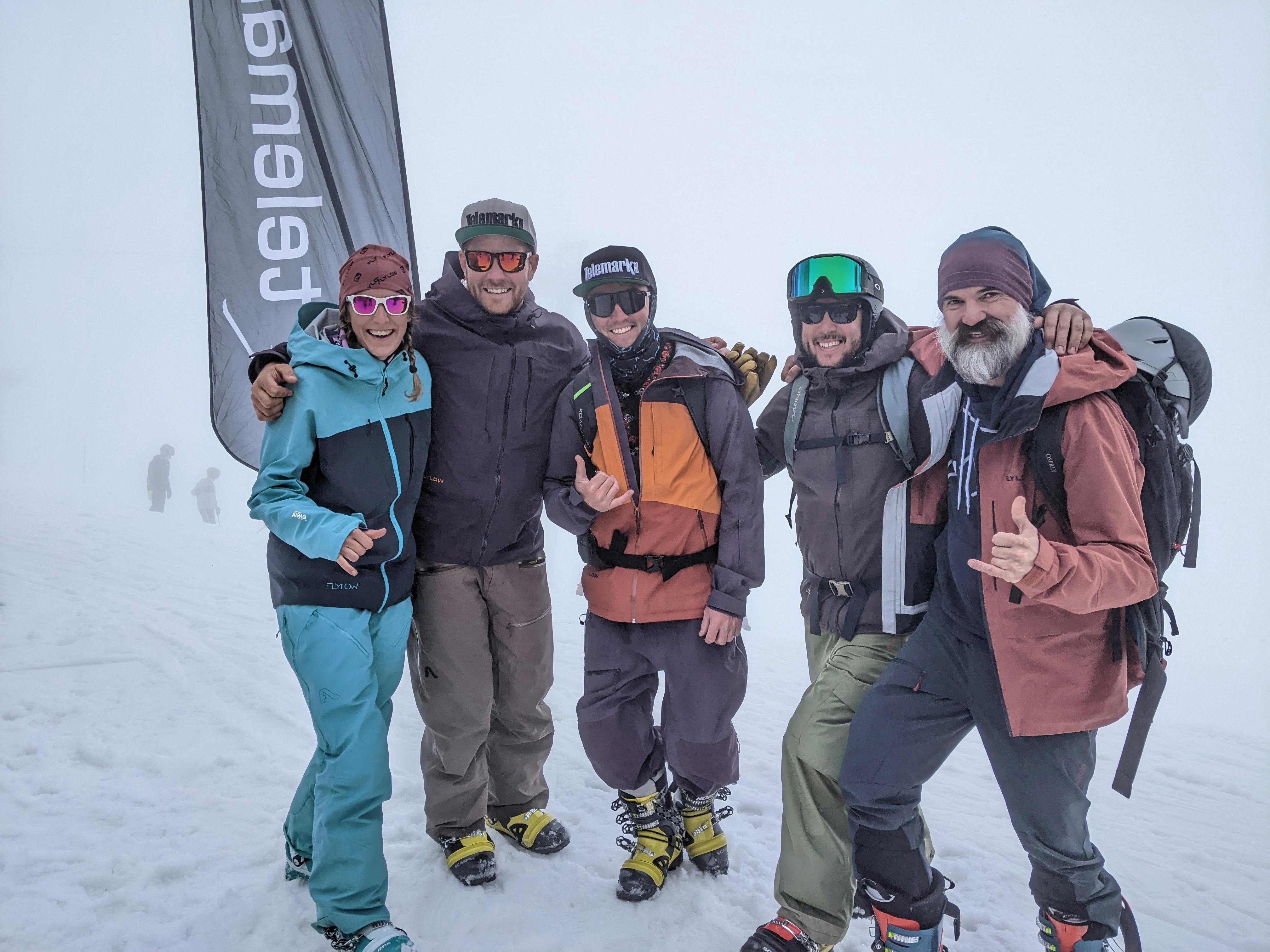 Five people wearing ski gear all hug each other and pose for a picture. There is snow on the ground. 