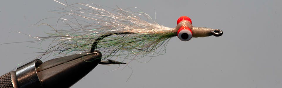 A Clouser Deep Minnow fly is held in a grip. Its eyes are orange and the material is green and silver. 