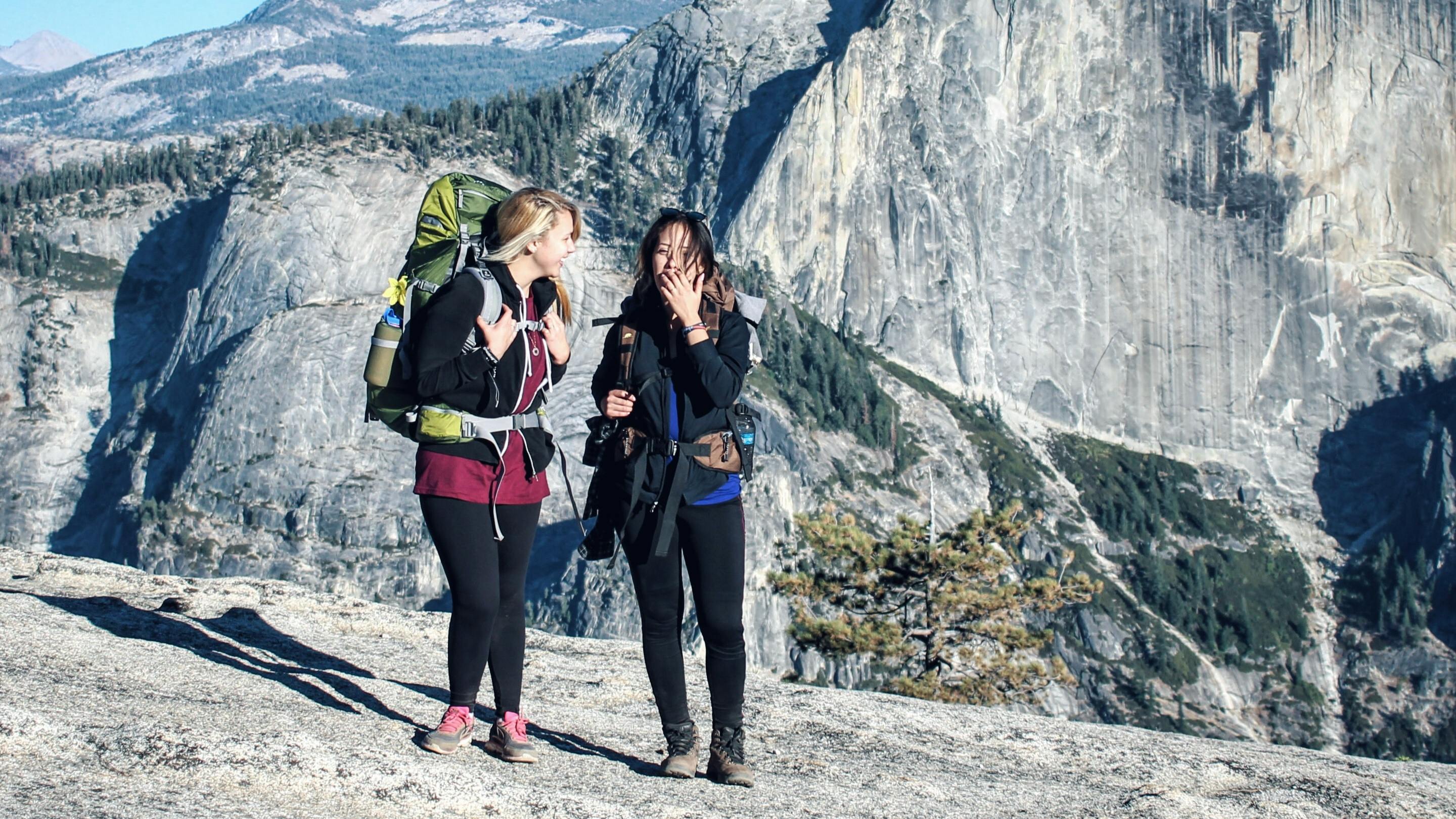 Two women with hiking backpacks on stand at the top of a hike. There are rocks and mountains behind them. 