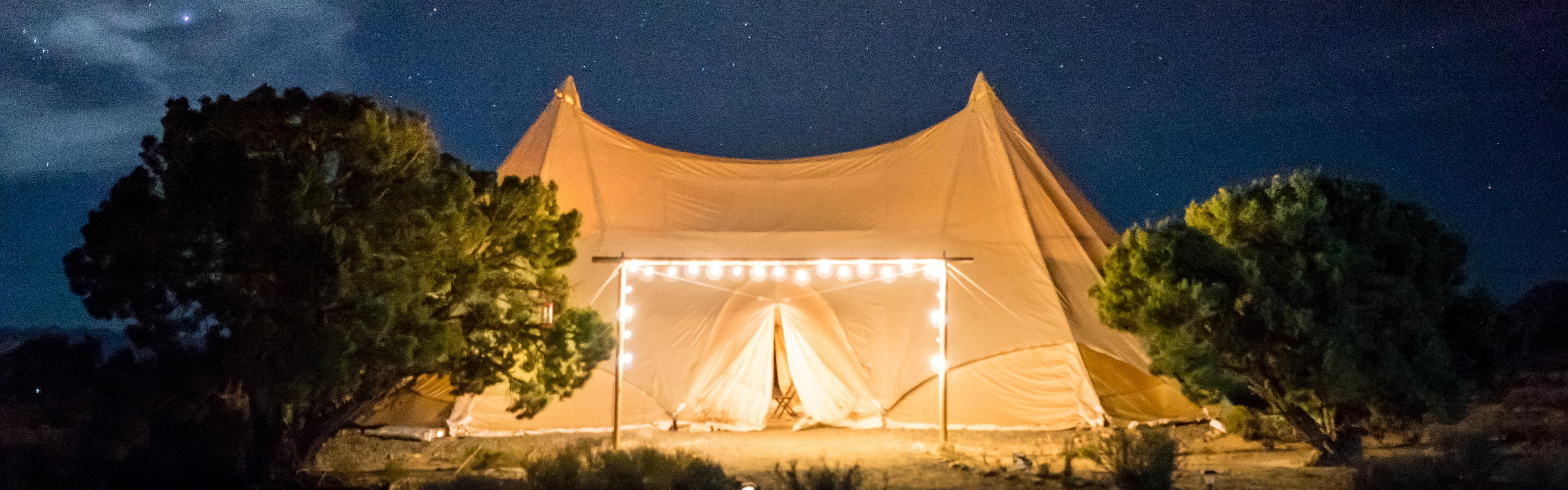 A large white tent is illuminated by a frame of lights. The night sky in the background is full of stars.