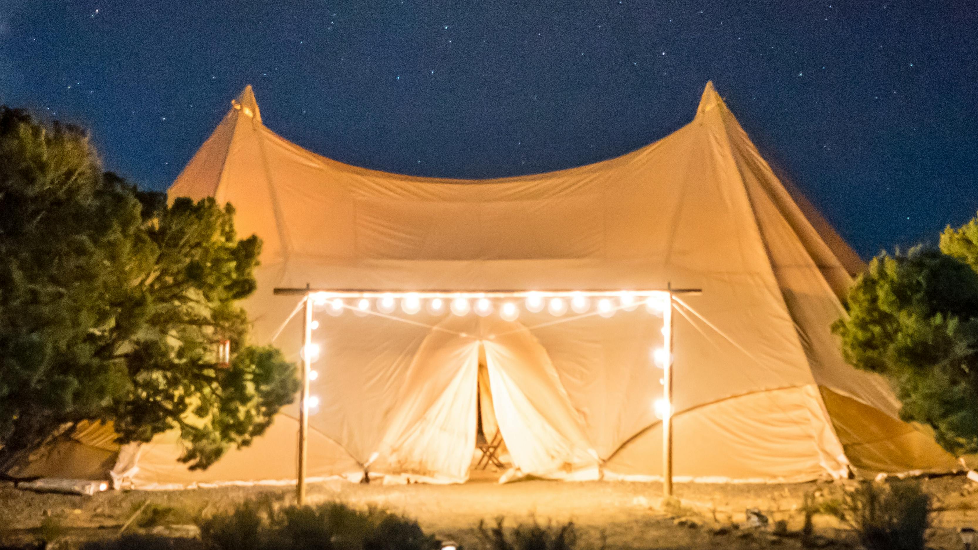 A large white tent is illuminated by a frame of lights. The night sky in the background is full of stars.