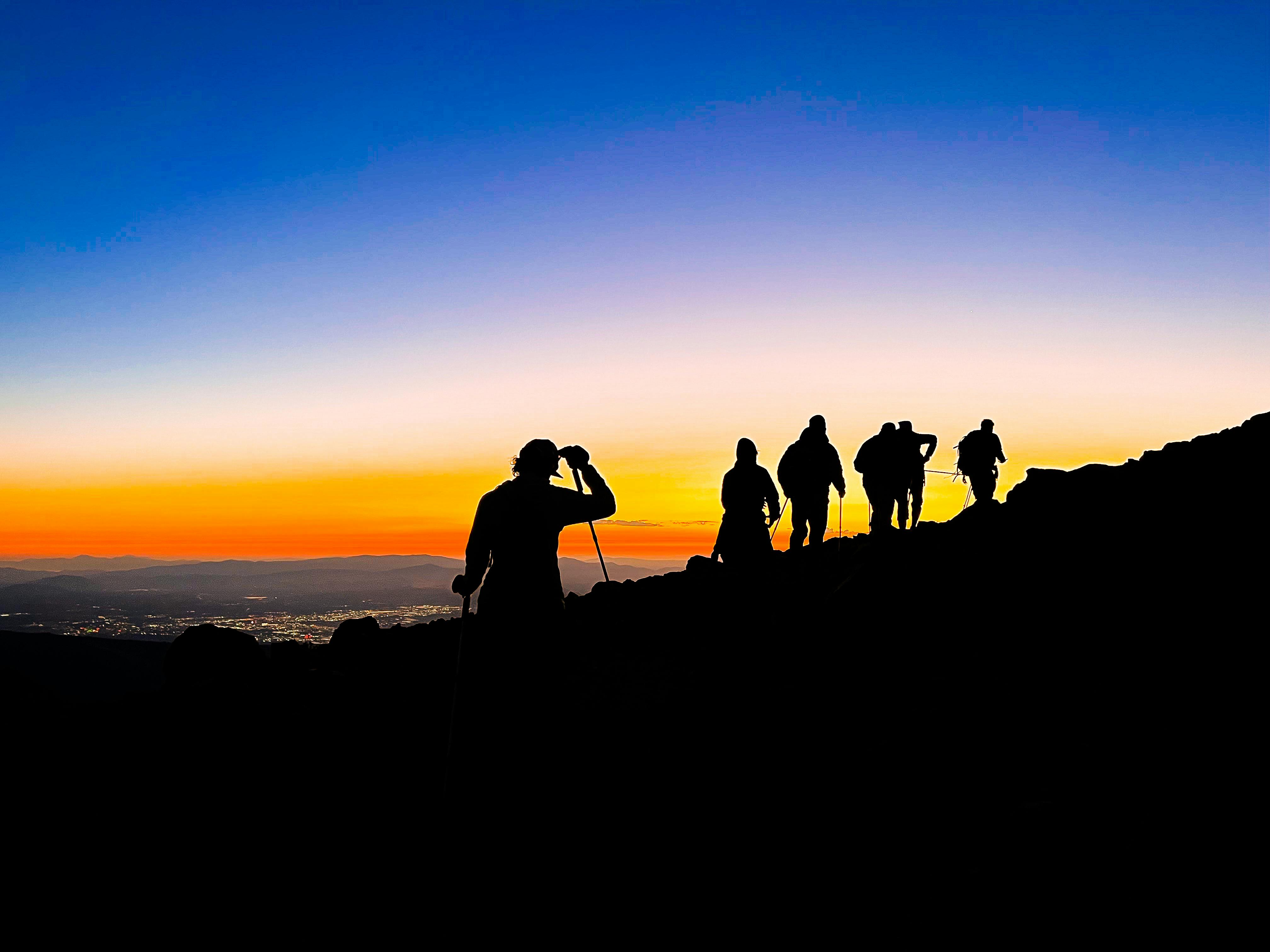A group of hikers walks along a ridge at sunset or sunrise with a sparkling city in the valley below them.