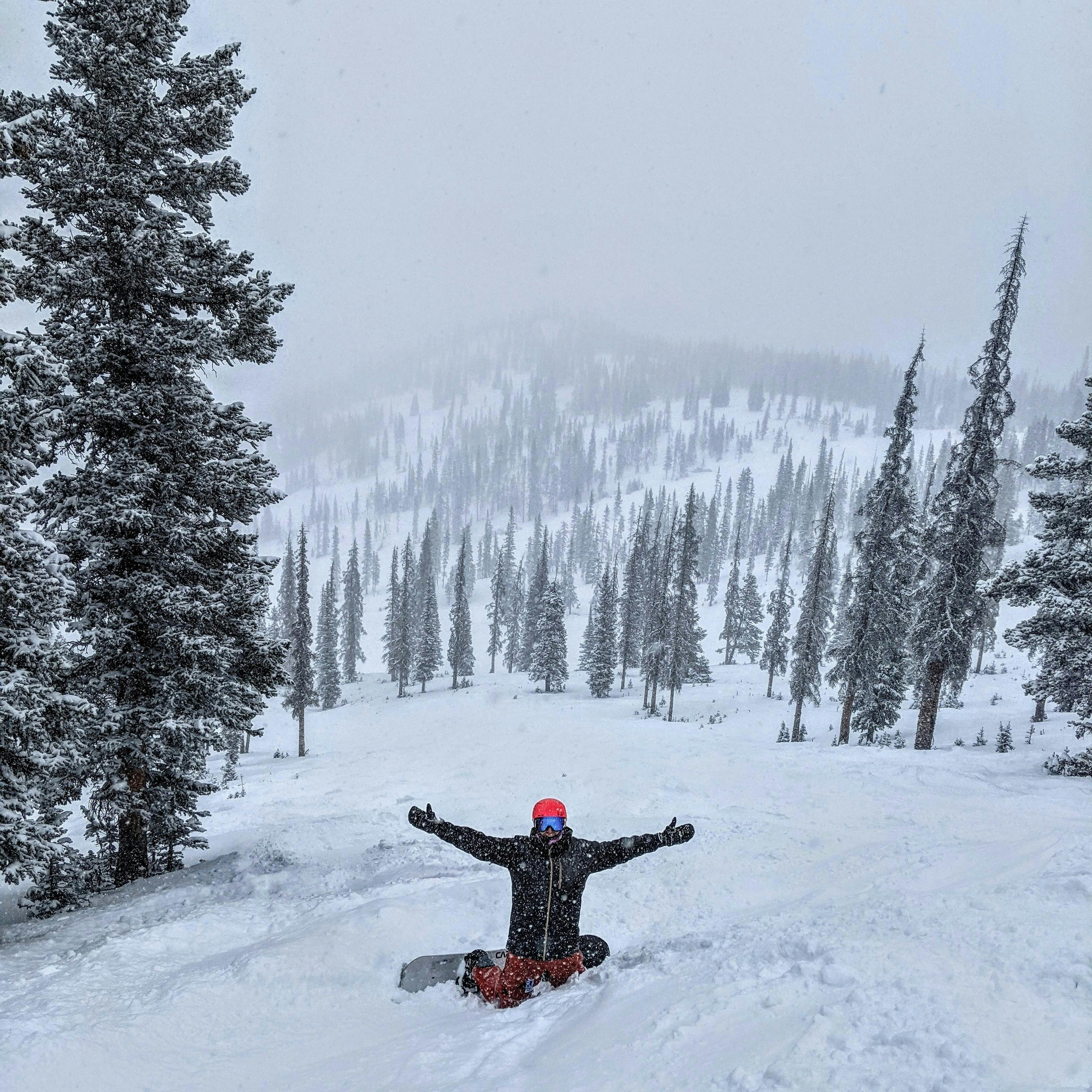 A snowboarder with his arms up kneeling in a snowy patch on a ski run. 
