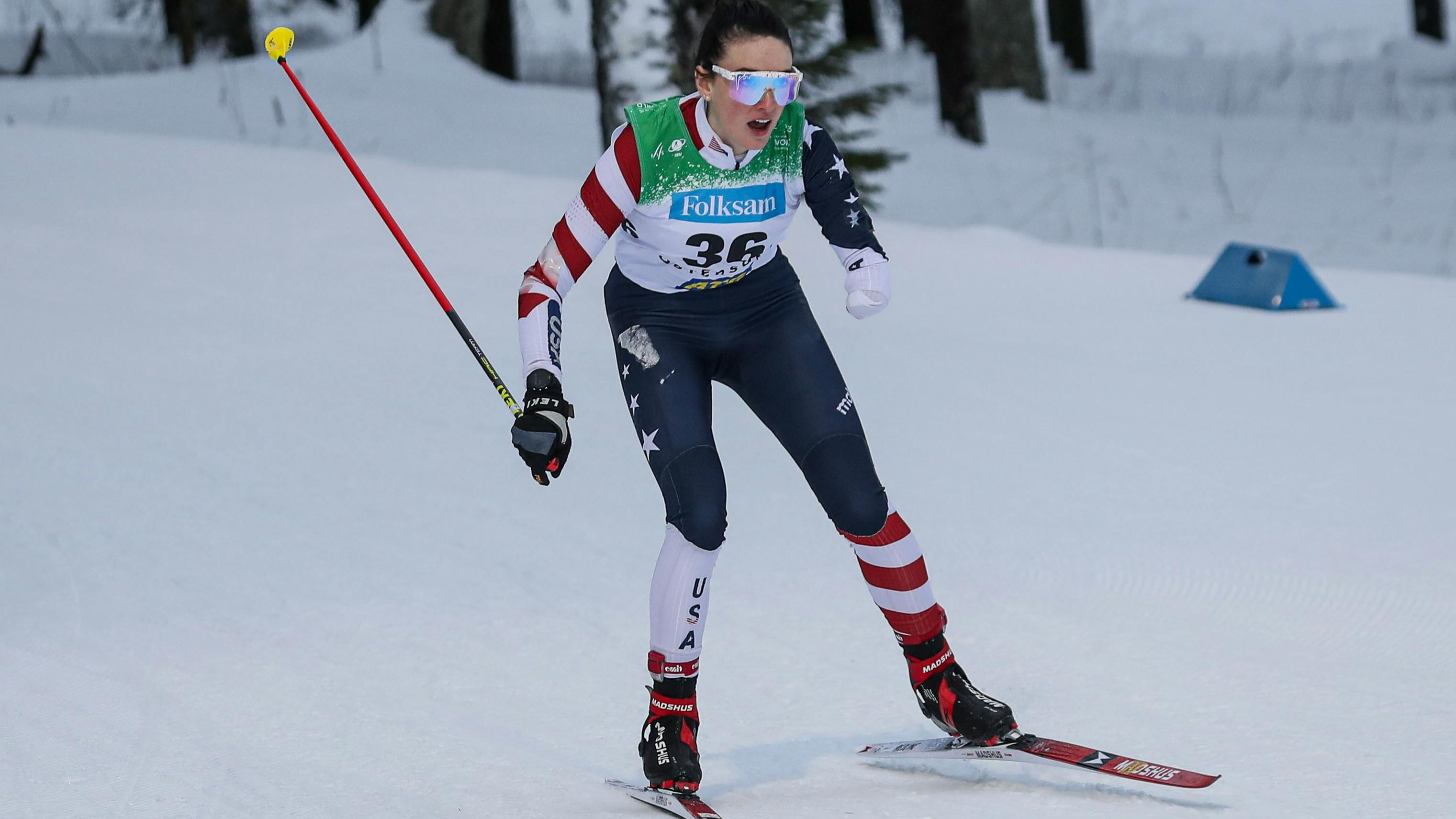 A nordic skier skis down a nordic track. She is wearing a speed suit that says "USA" and has an American flag print. She is only using one pole and is missing her left hand and forearm. 